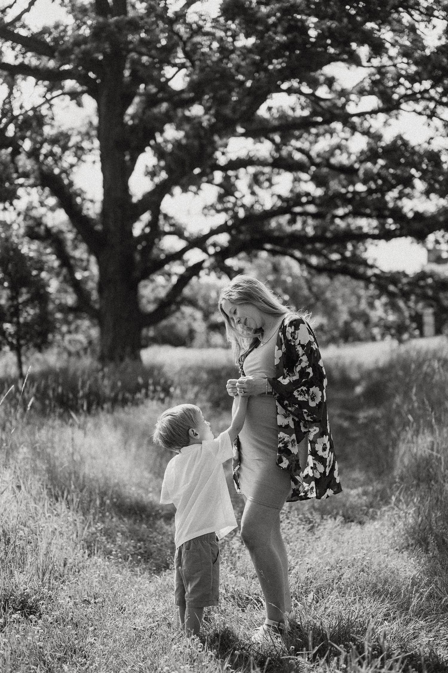 Black and white photo of a pregnant woman holding hands with her young son in a grassy field with a large tree.