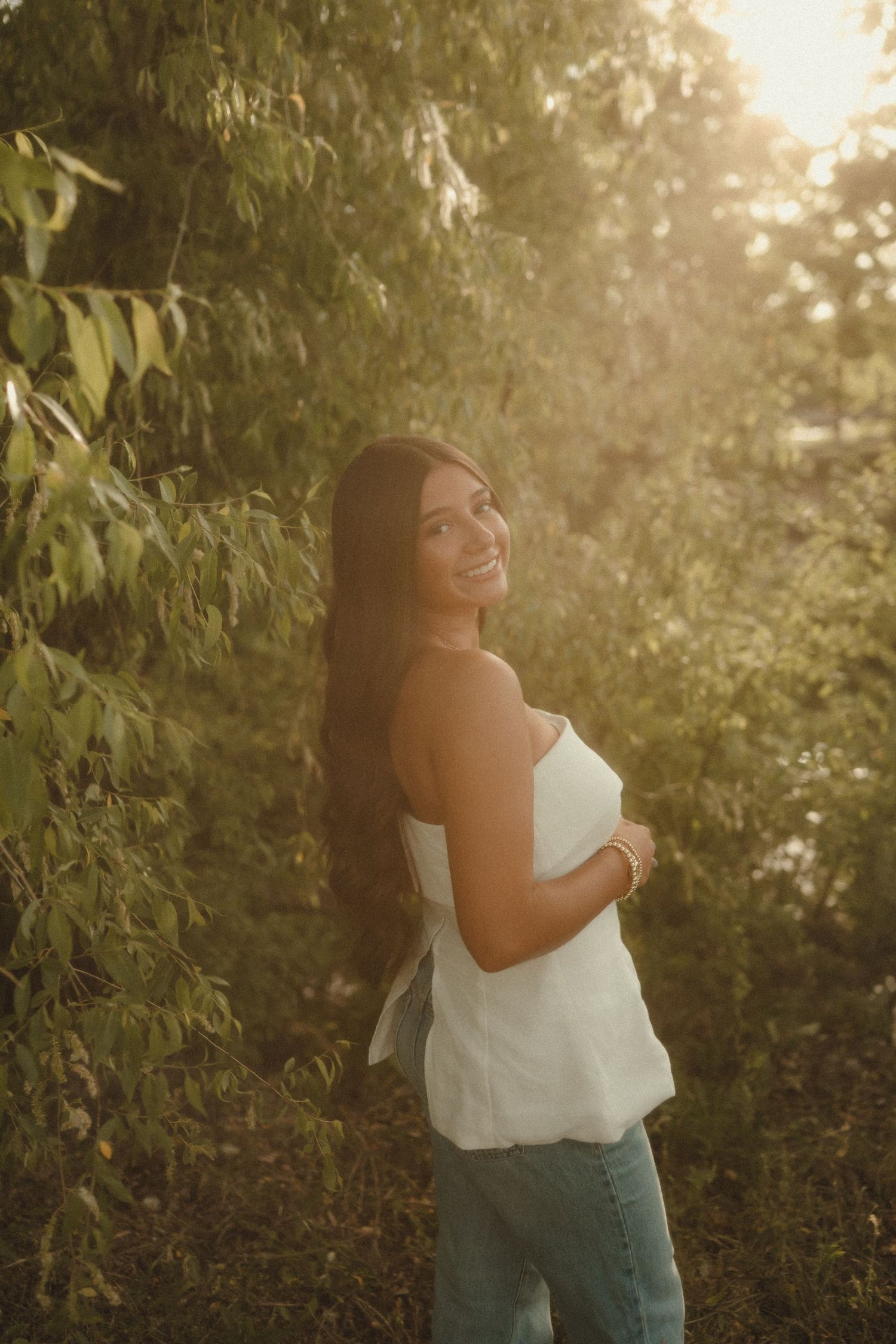Smiling young woman in white top and jeans standing among green trees in golden sunlight.