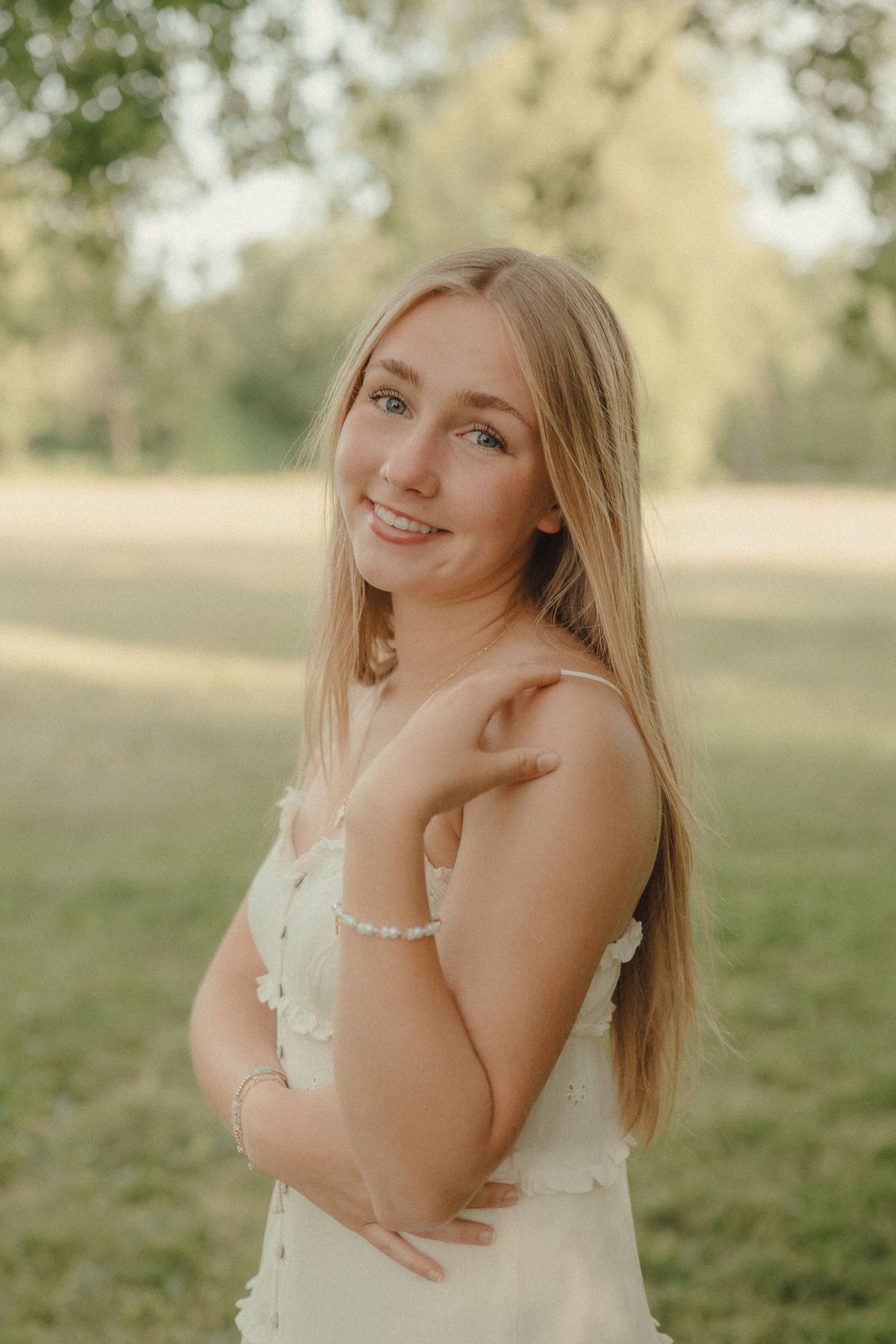 Young blonde woman in white floral dress smiling outdoors in a sunny green park setting.
