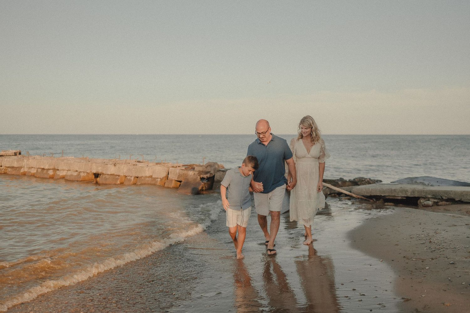 Family of three walking along a sandy beach at sunset, waves gently lapping the shore.