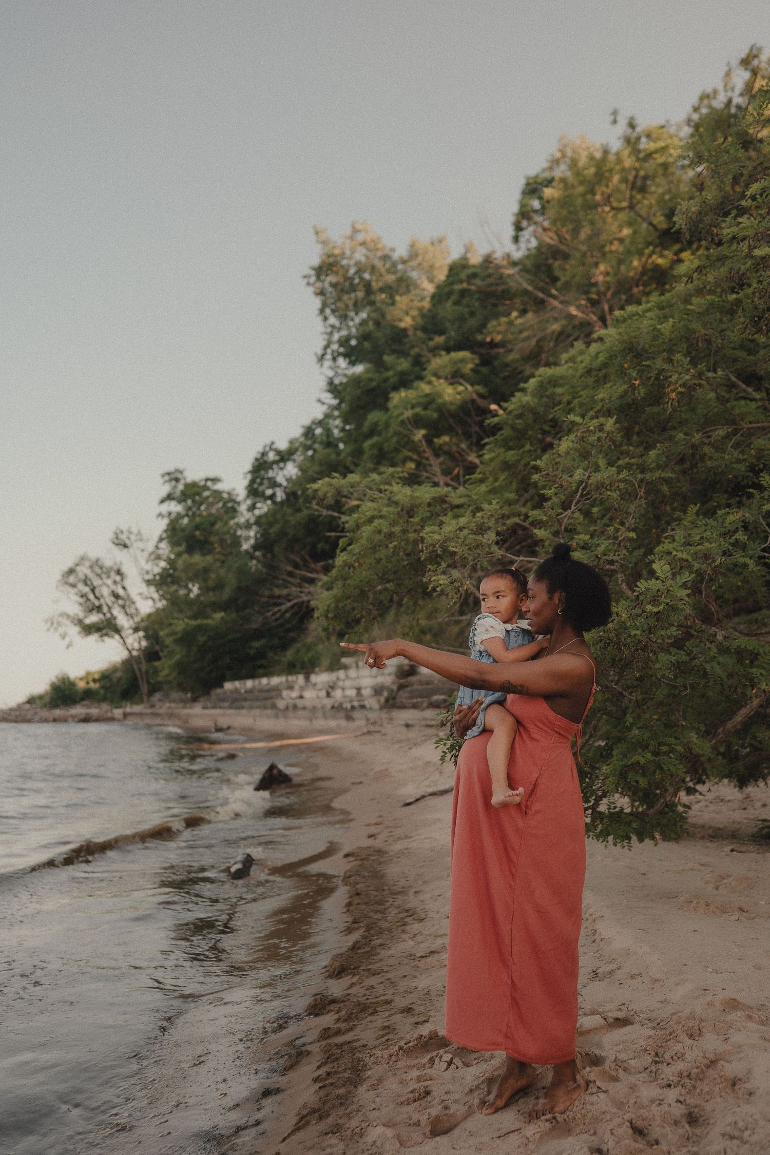 A mother in a coral dress holds her baby on a sandy beach, pointing toward the water at sunset.
