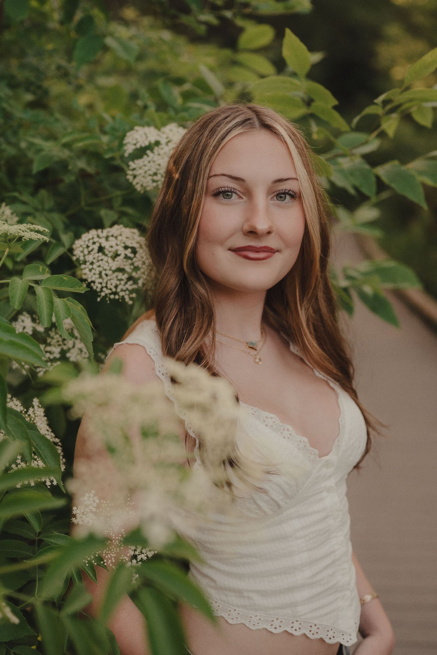Young woman with long hair smiling among white flowering elderberry bushes in a white lace top.