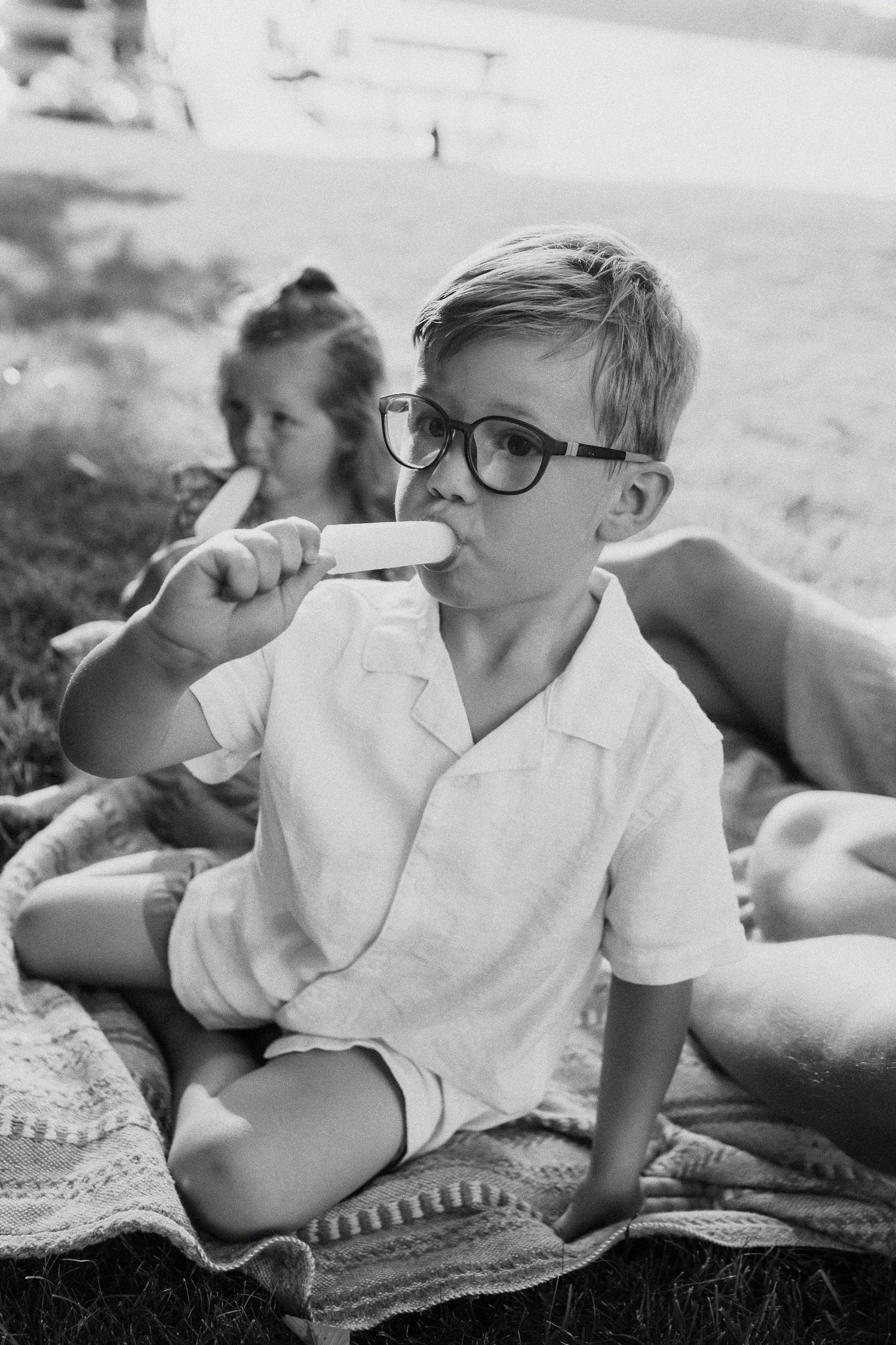 Black and white photo of a boy with glasses eating a popsicle beside a girl on a blanket outdoors.