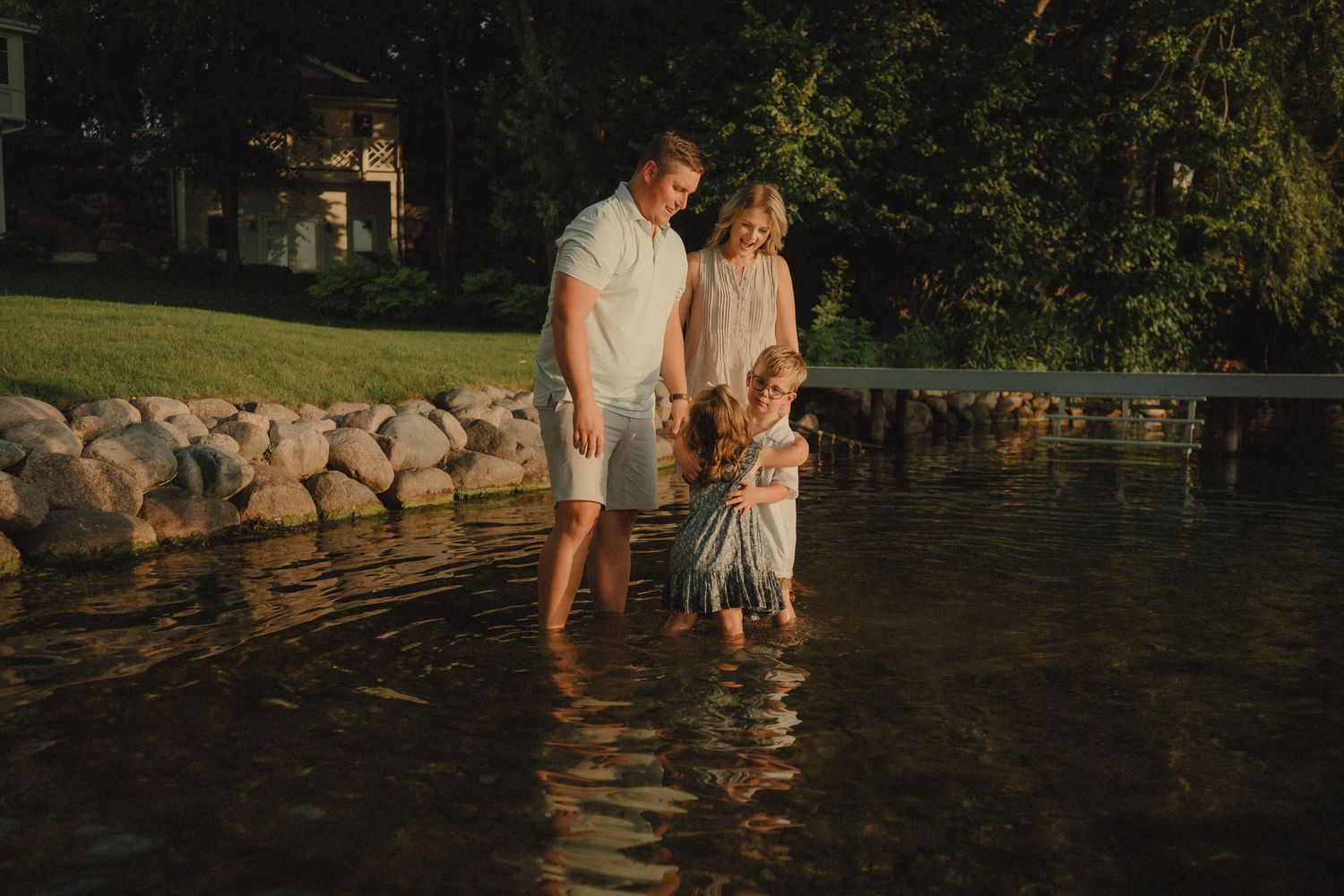 Family of three wading in shallow water near a rocky shoreline at sunset in a serene outdoor setting.