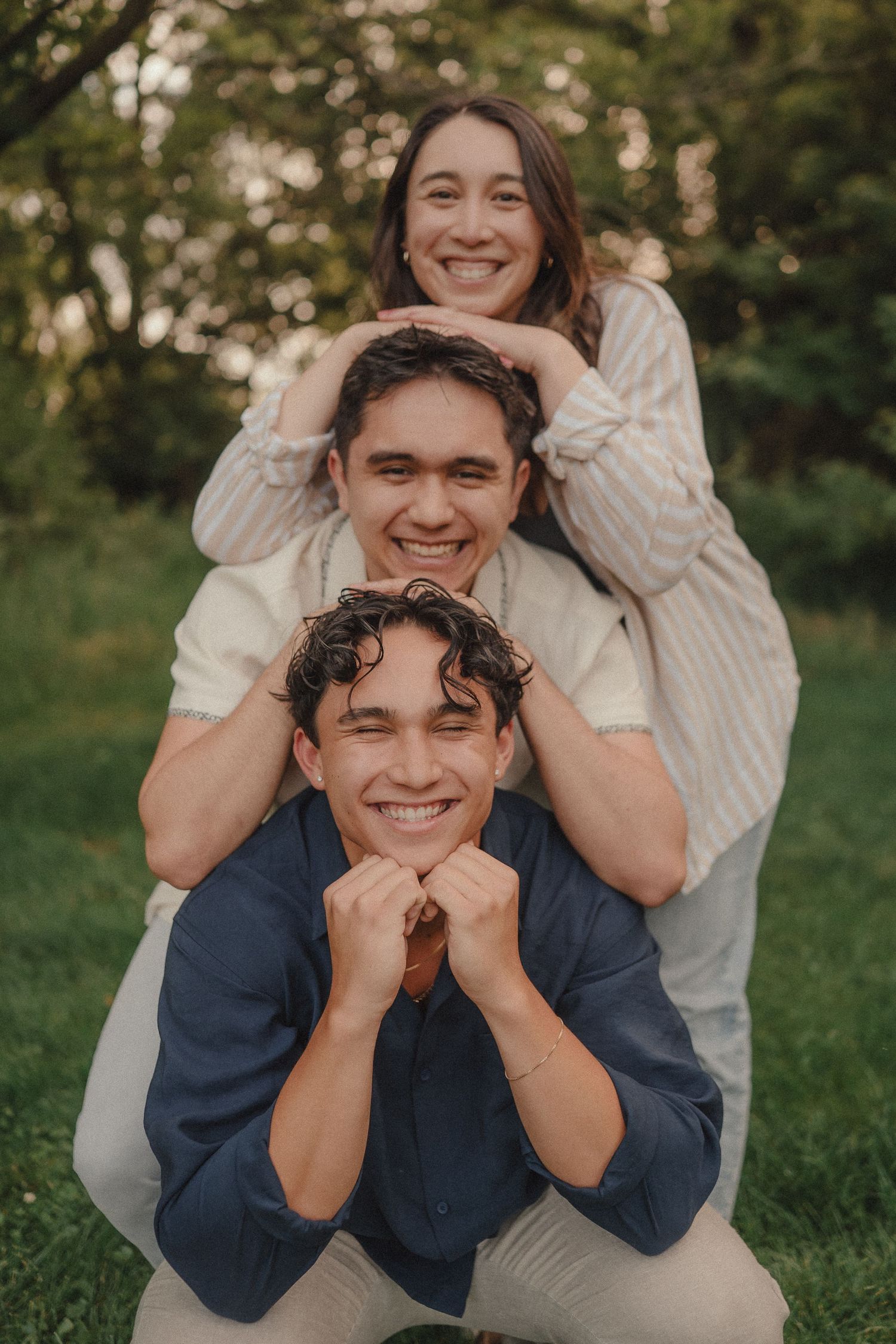 Three smiling young people pose playfully outdoors, stacked vertically in a park setting with green trees behind them.