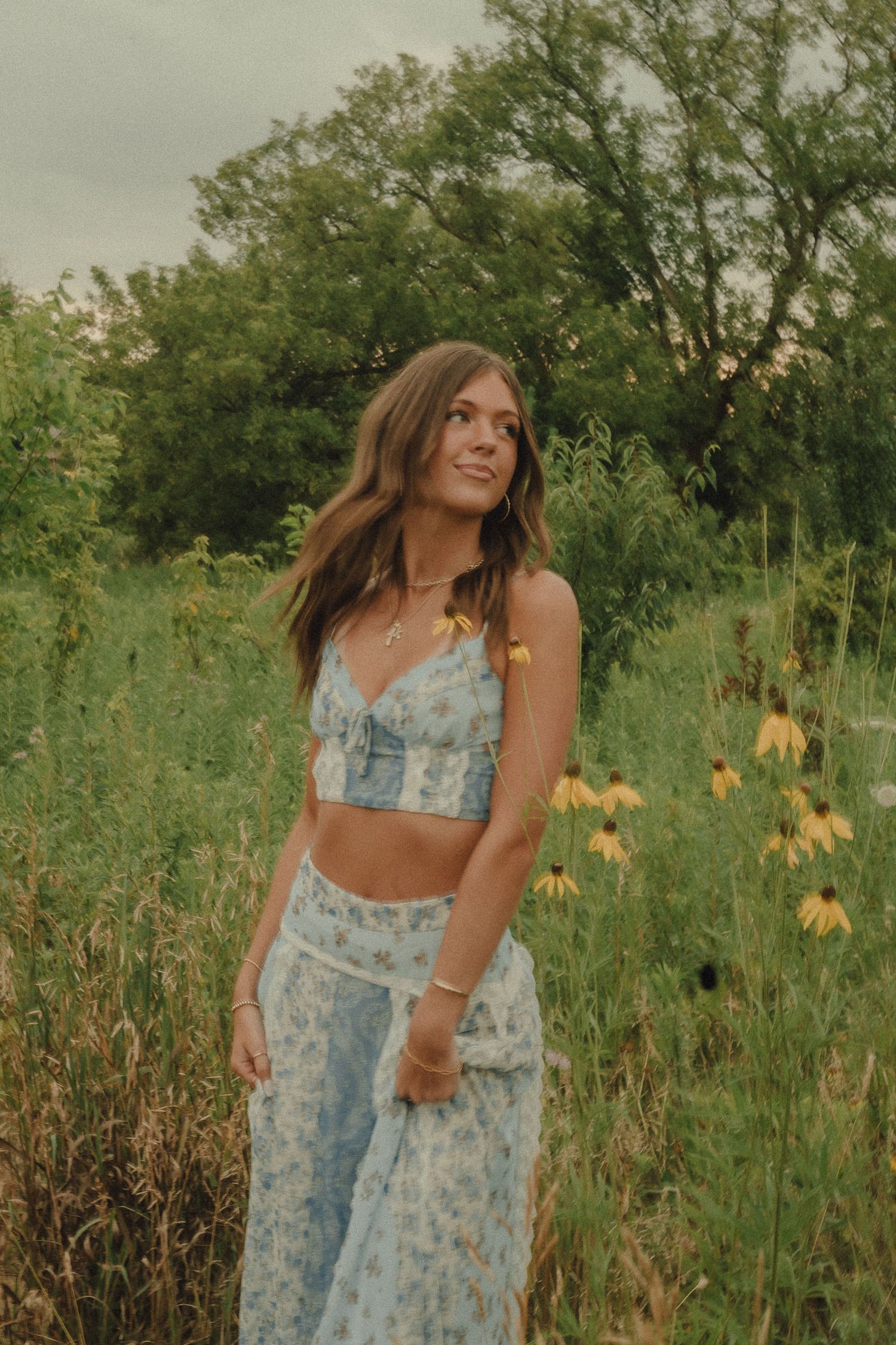 Smiling young woman in blue floral crop top and skirt standing among yellow wildflowers in a green field.