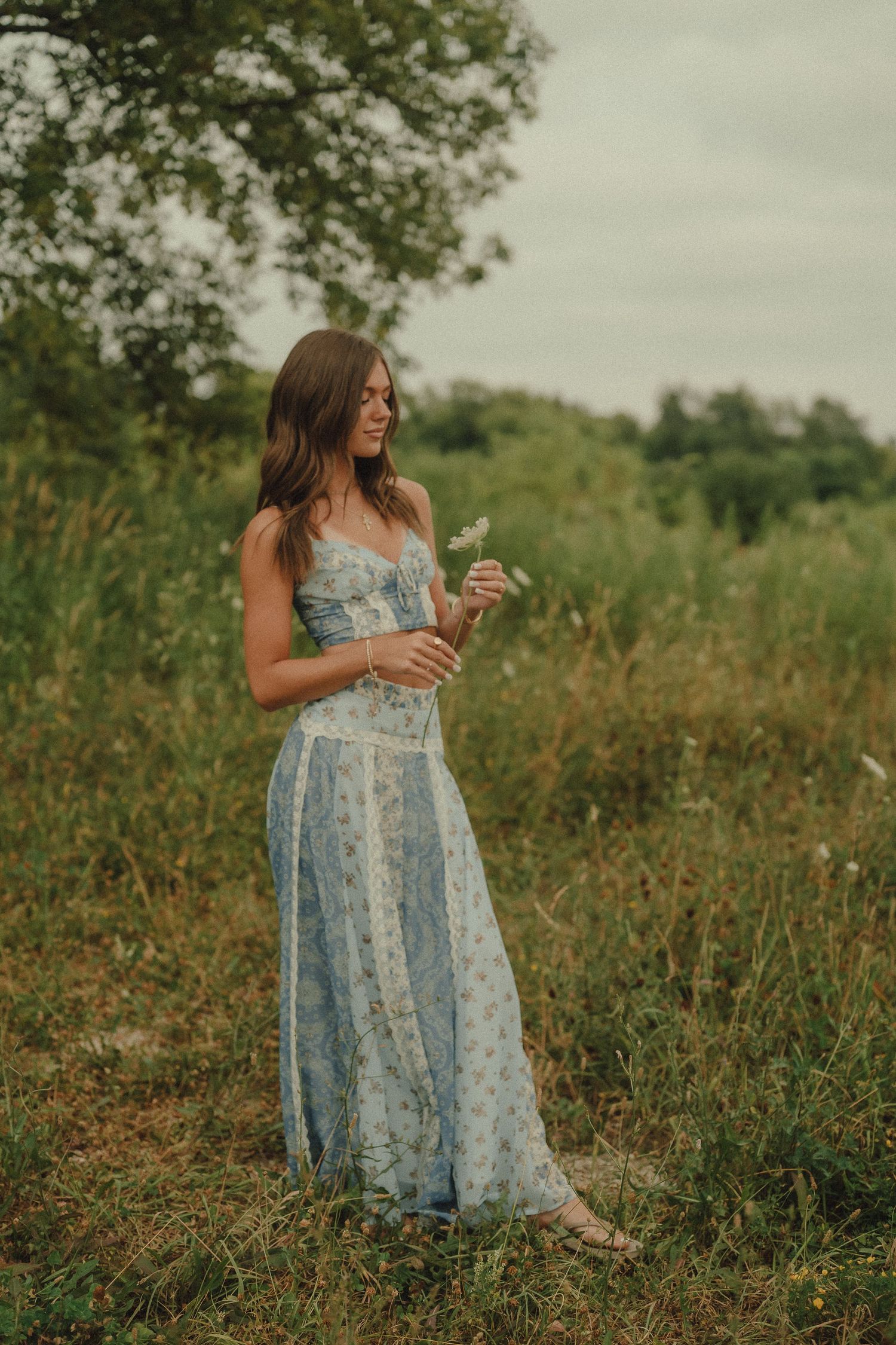 Young woman in blue floral maxi dress holding wildflowers in a lush green field.