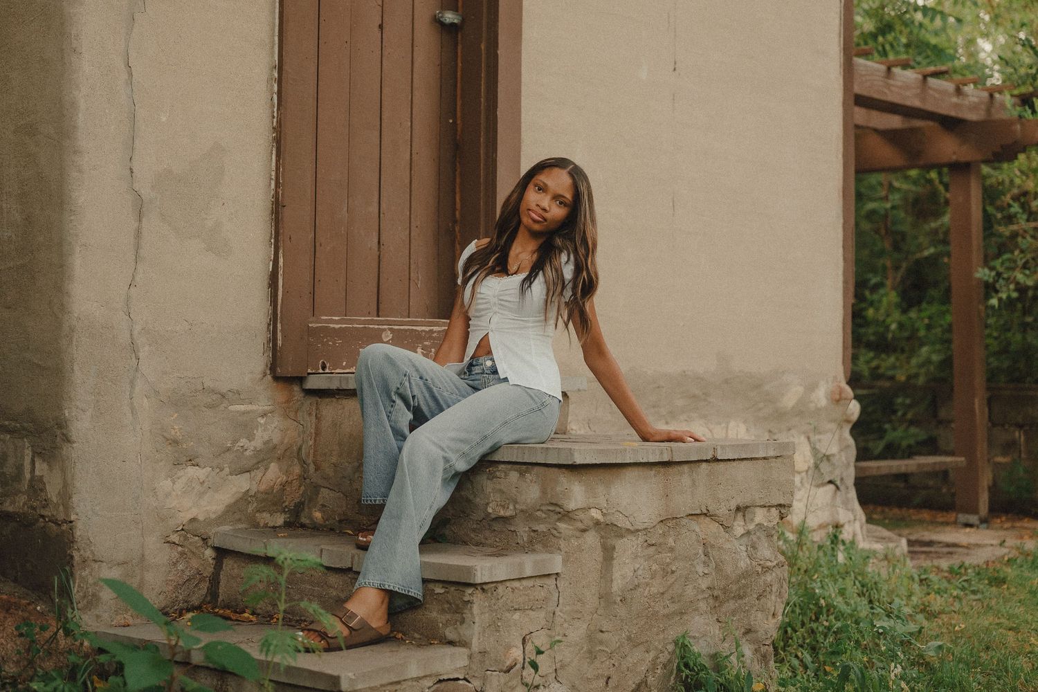 Young woman in white top and jeans sitting on stone steps outside a rustic building.