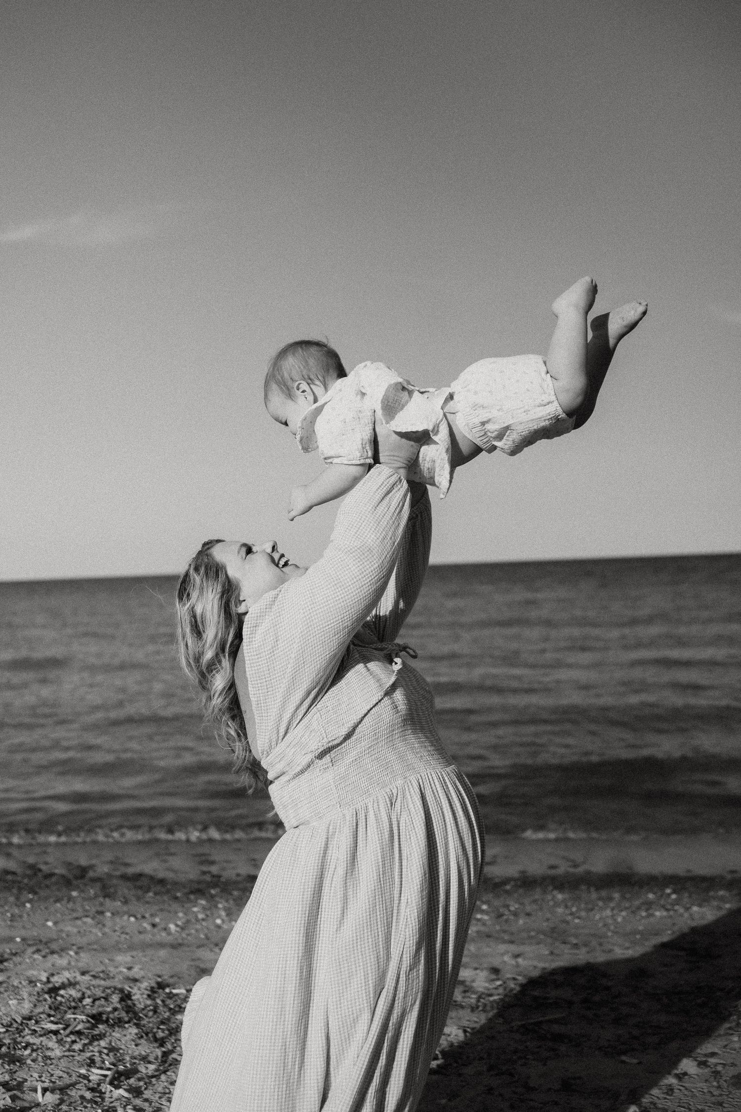 Black and white photo of mother lifting baby in air on rocky beach with ocean behind them.