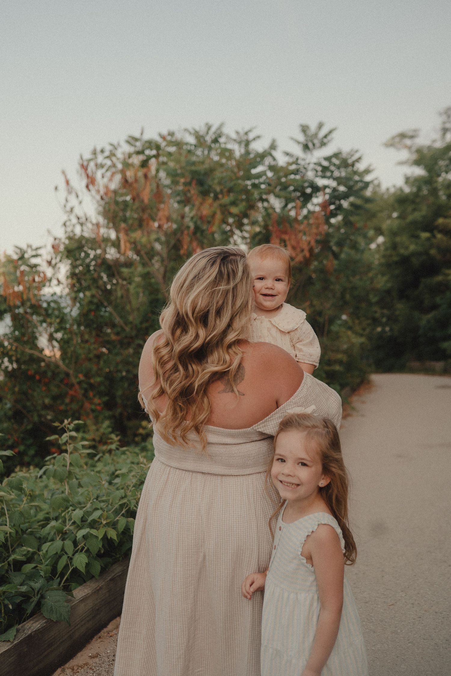 A mother holds a baby on her shoulder while her young daughter smiles beside her outdoors.