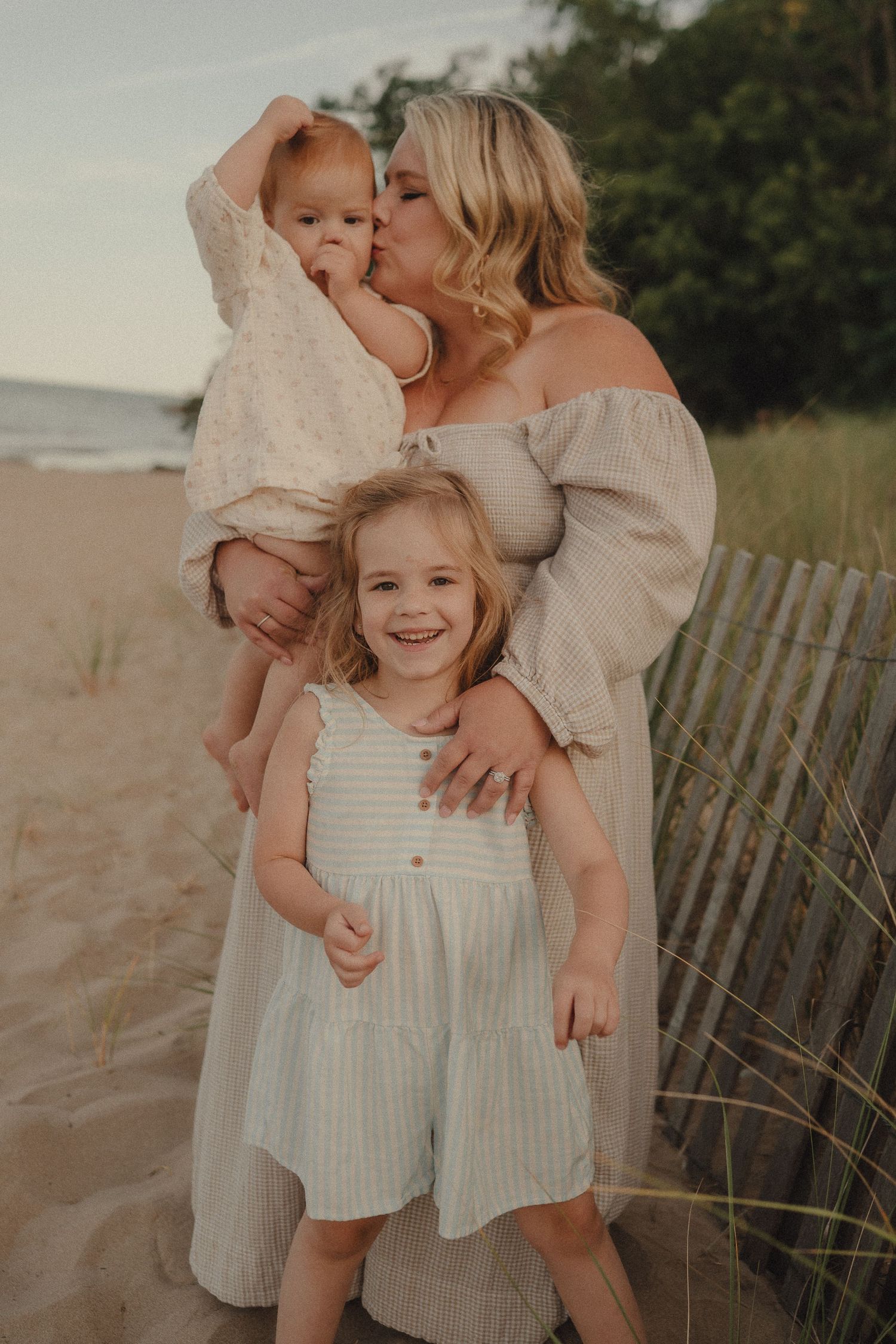 Loving mother kissing red-haired baby while smiling toddler girl stands in front on beach.