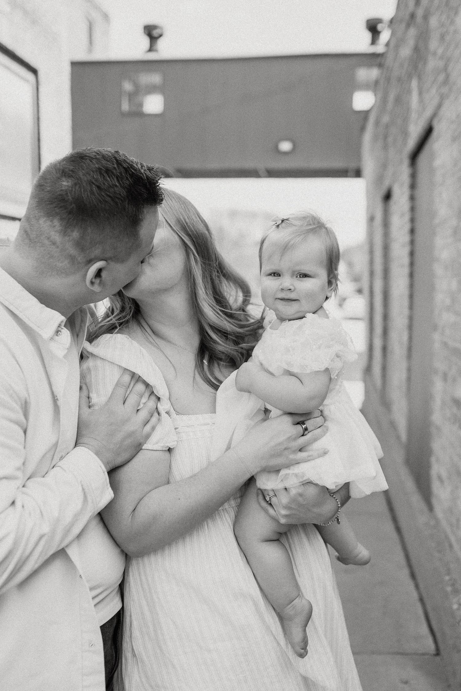 Black and white photo of a father kissing mother on cheek while she holds their smiling baby girl outdoors.