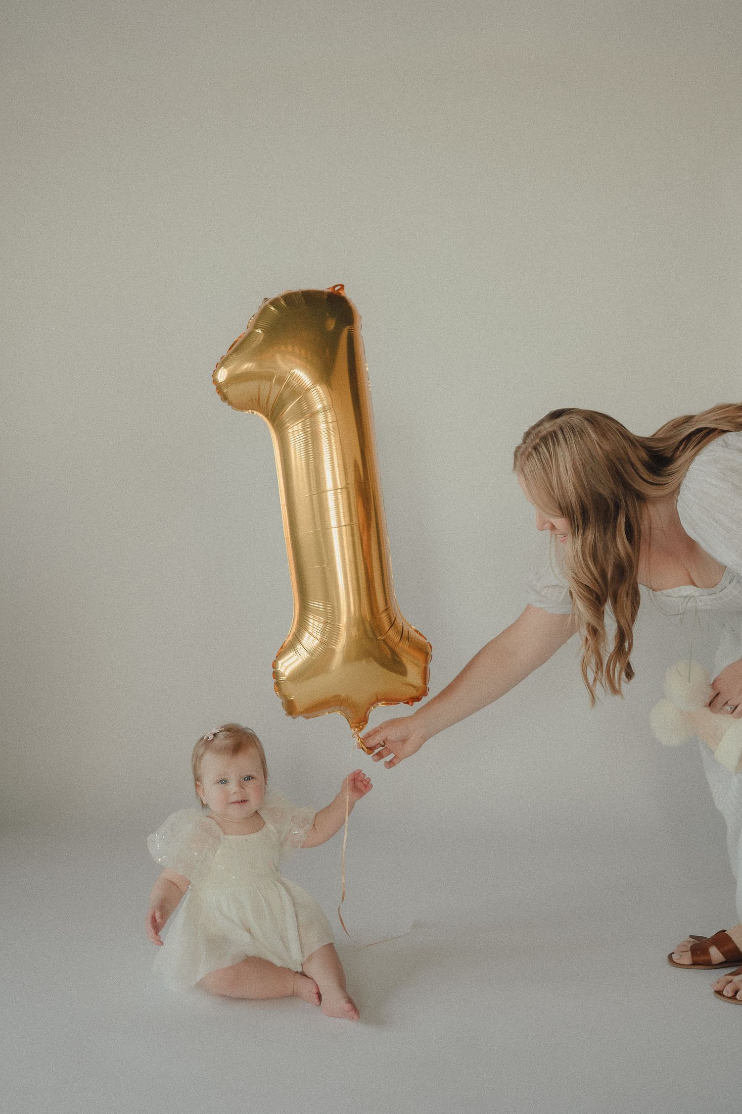 Baby girl in white dress holds gold number 1 balloon with older sister during first birthday photo session.