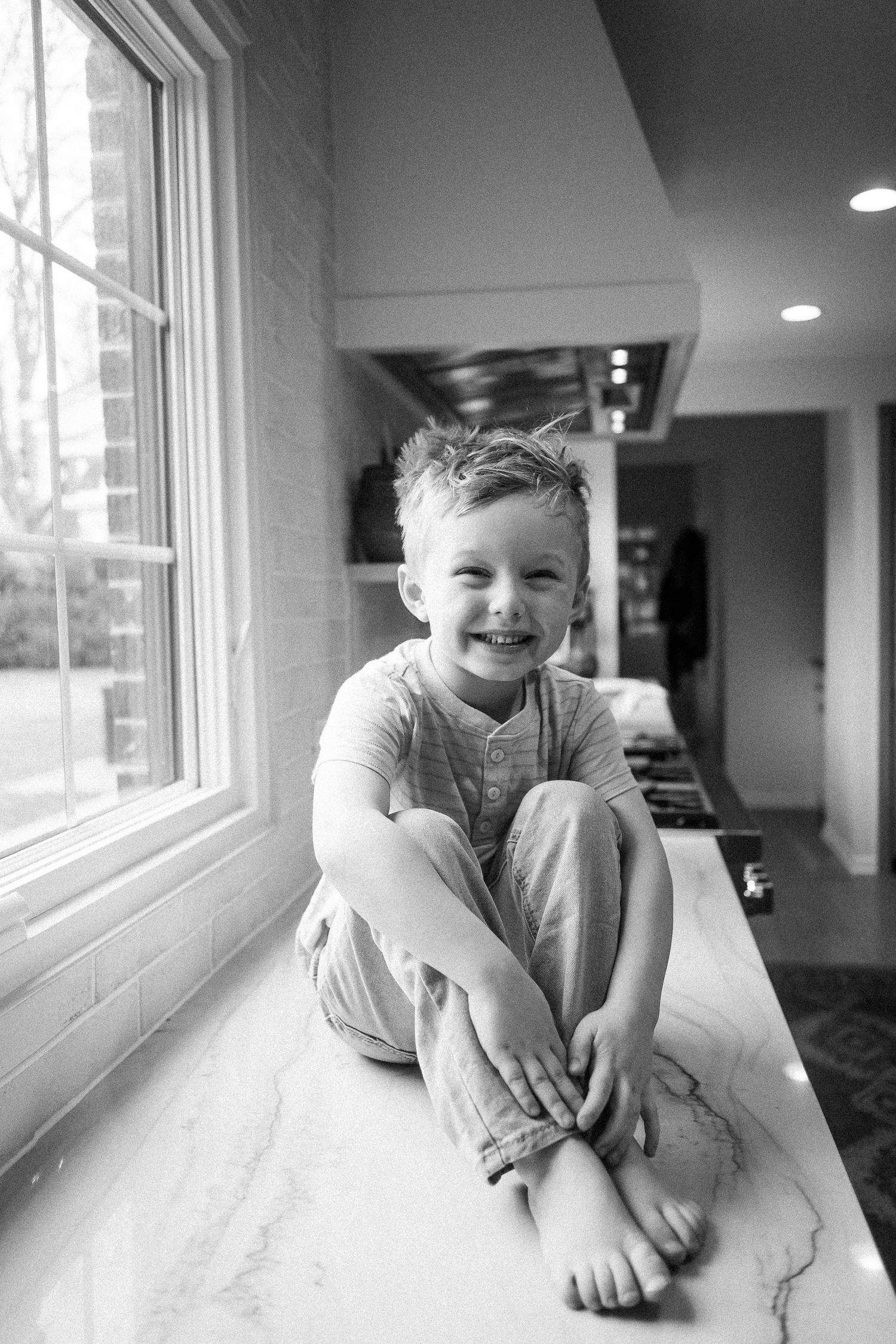 Black and white portrait of a smiling boy sitting cross-legged on a marble kitchen counter.