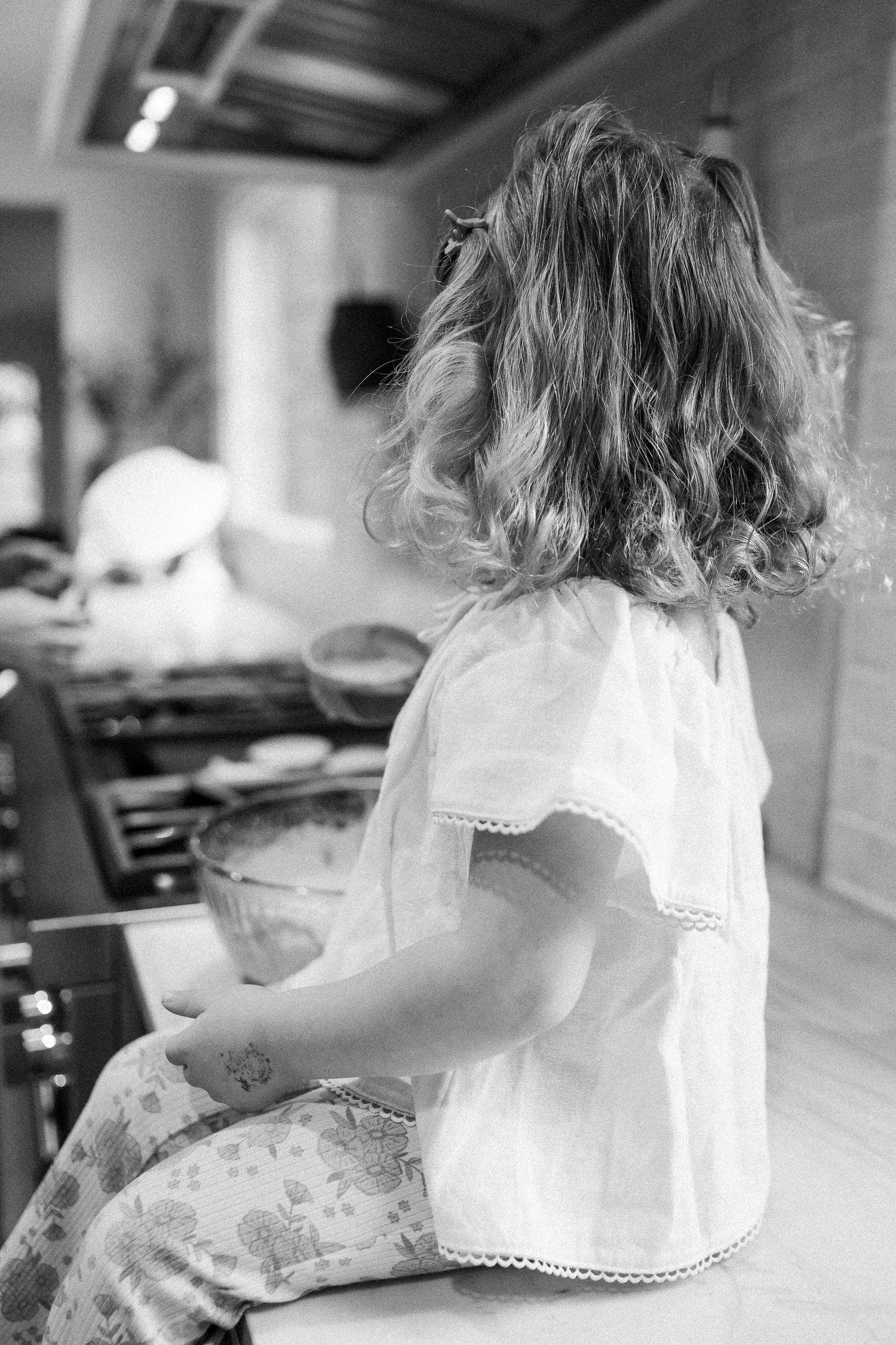 A young girl with curly hair sits at a kitchen counter in a white dress in black and white photo.