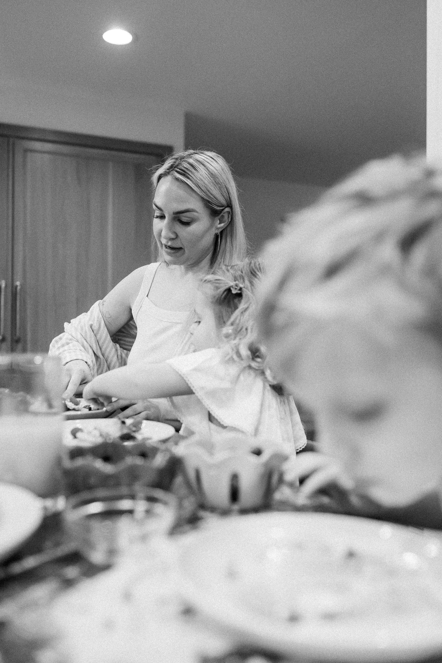 A blonde woman prepares food at a kitchen counter while a child watches in black and white photo.