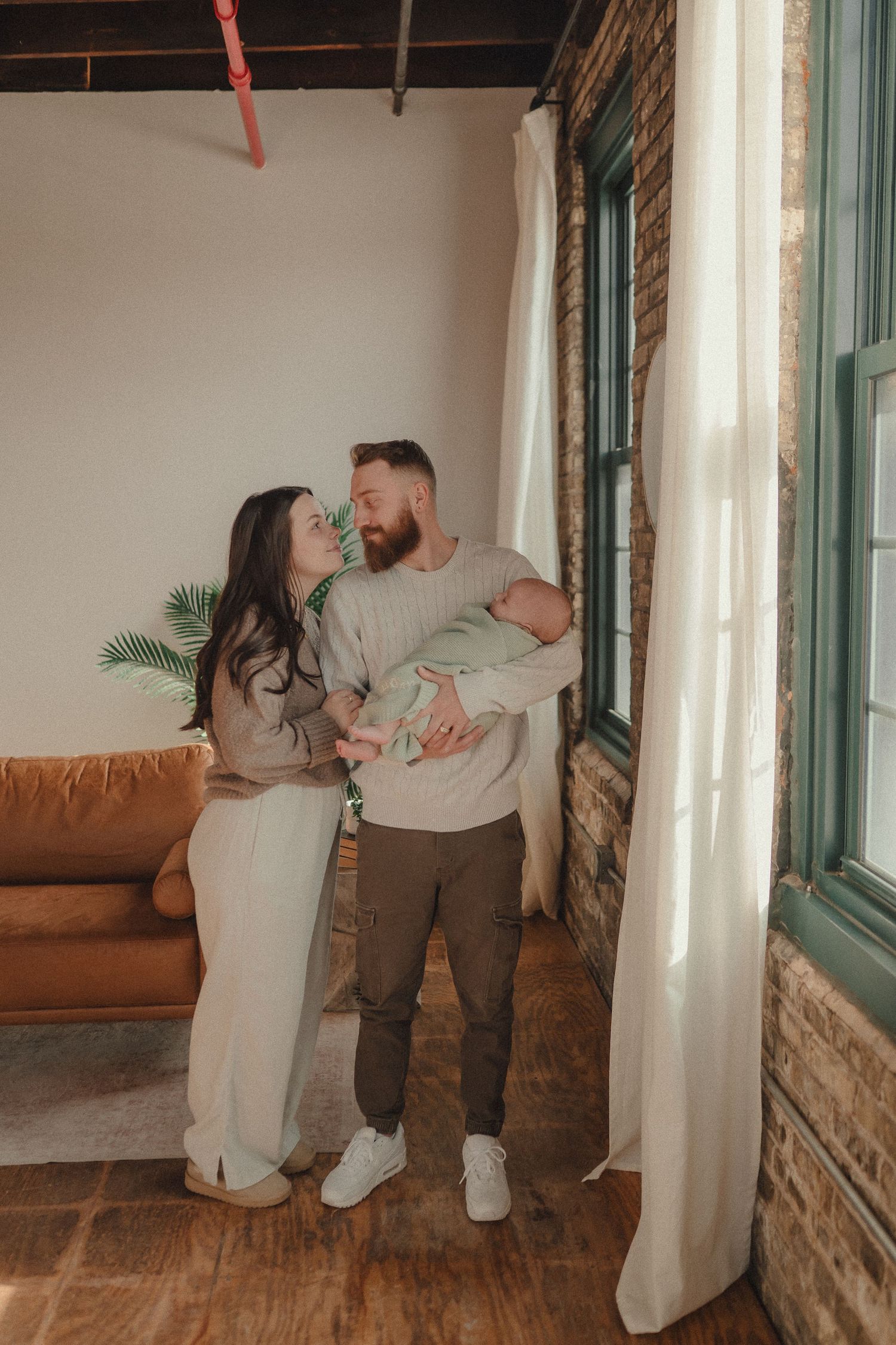 A loving family moment captured in front of large windows in an industrial-style space with brick walls.