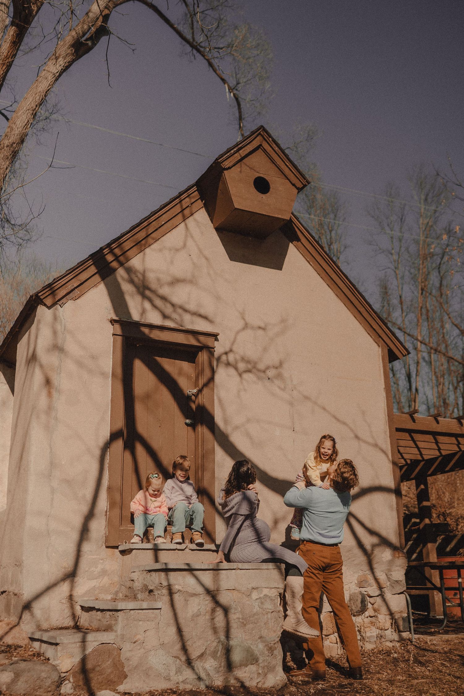 A family gathers on the steps of a small beige building with a birdhouse on top in warm sunlight.