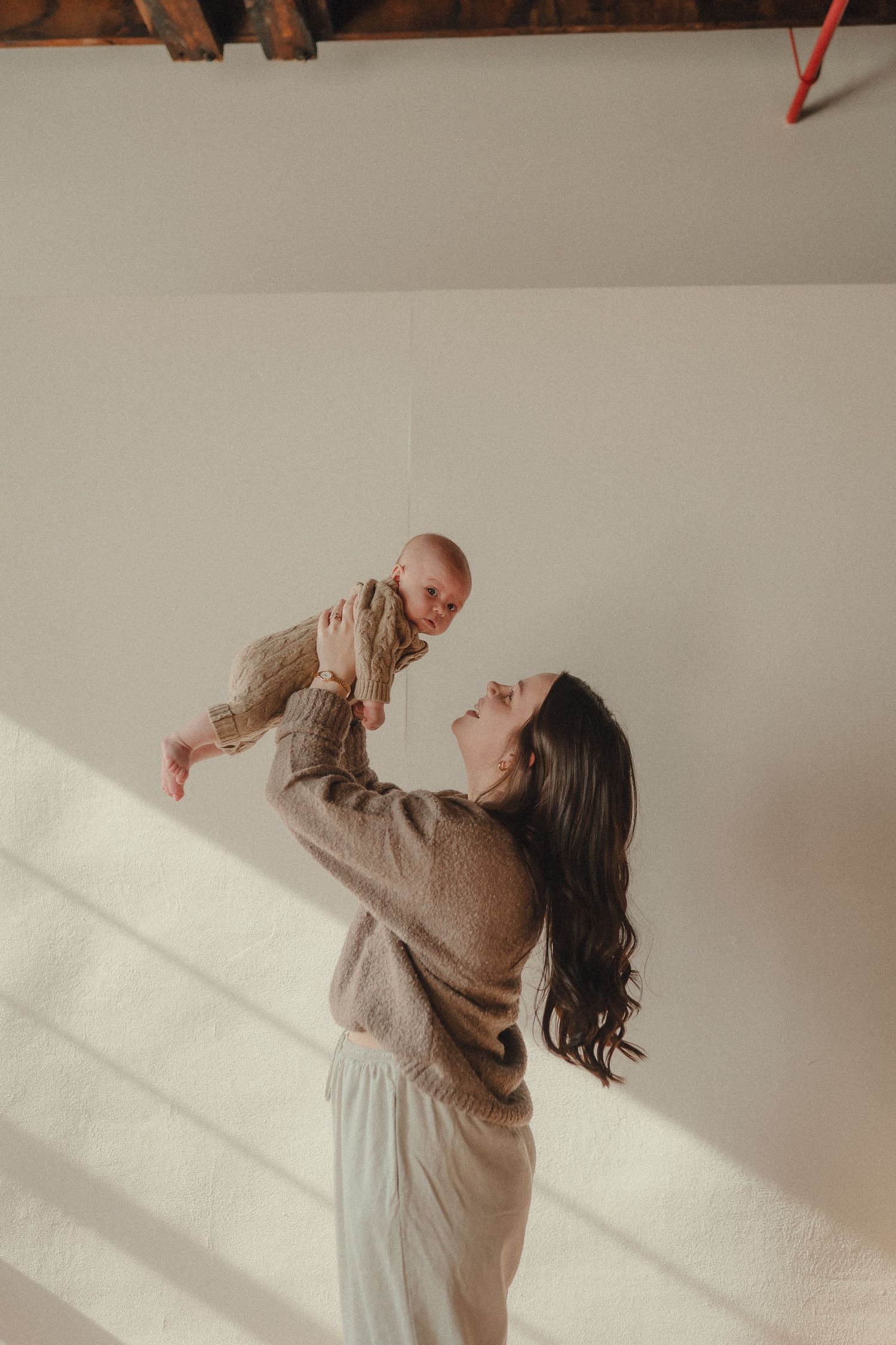 Woman in beige sweater posing against white wall with dramatic natural lighting.