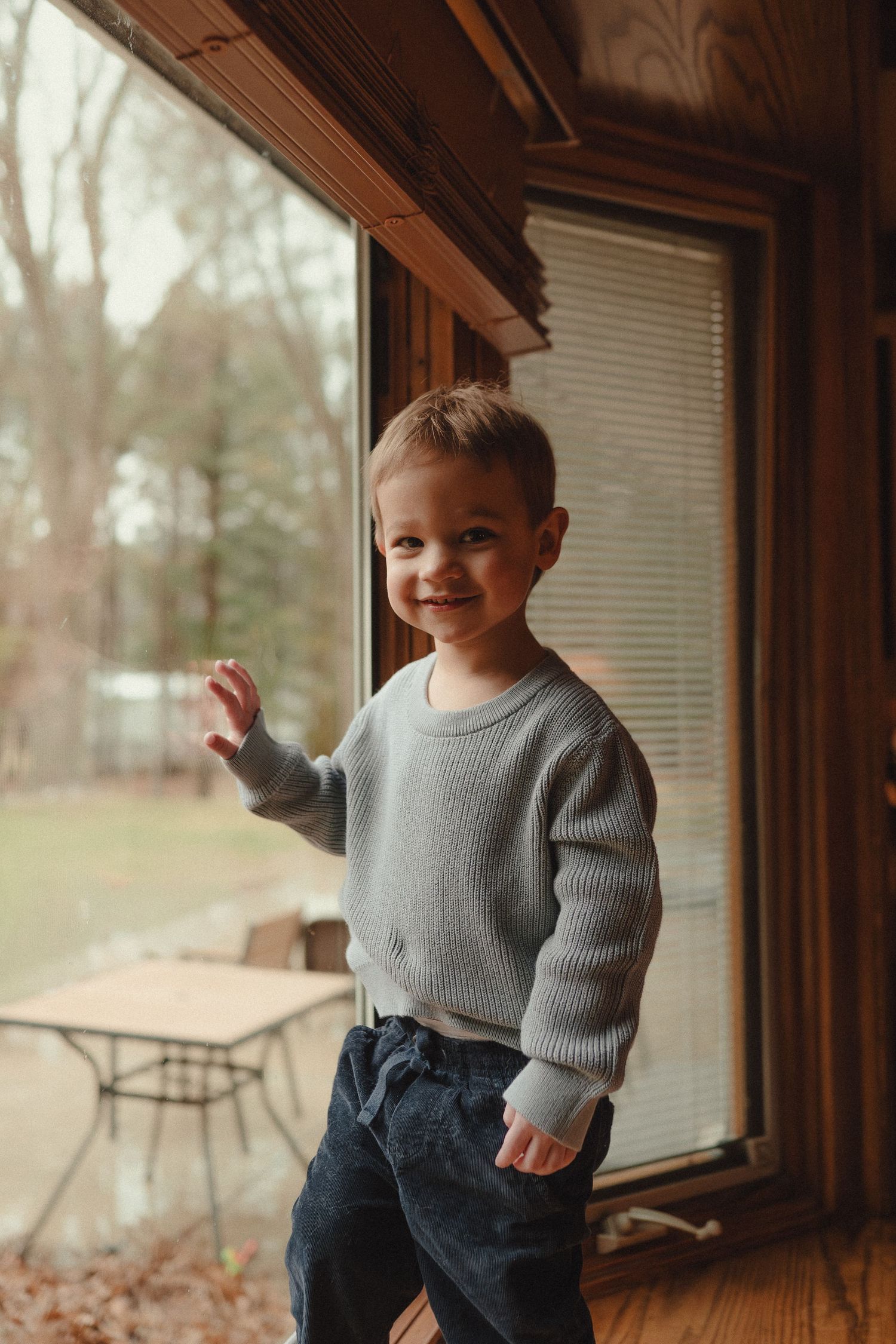 Child wearing a grey sweater and jeans stands by a window in warm natural light.