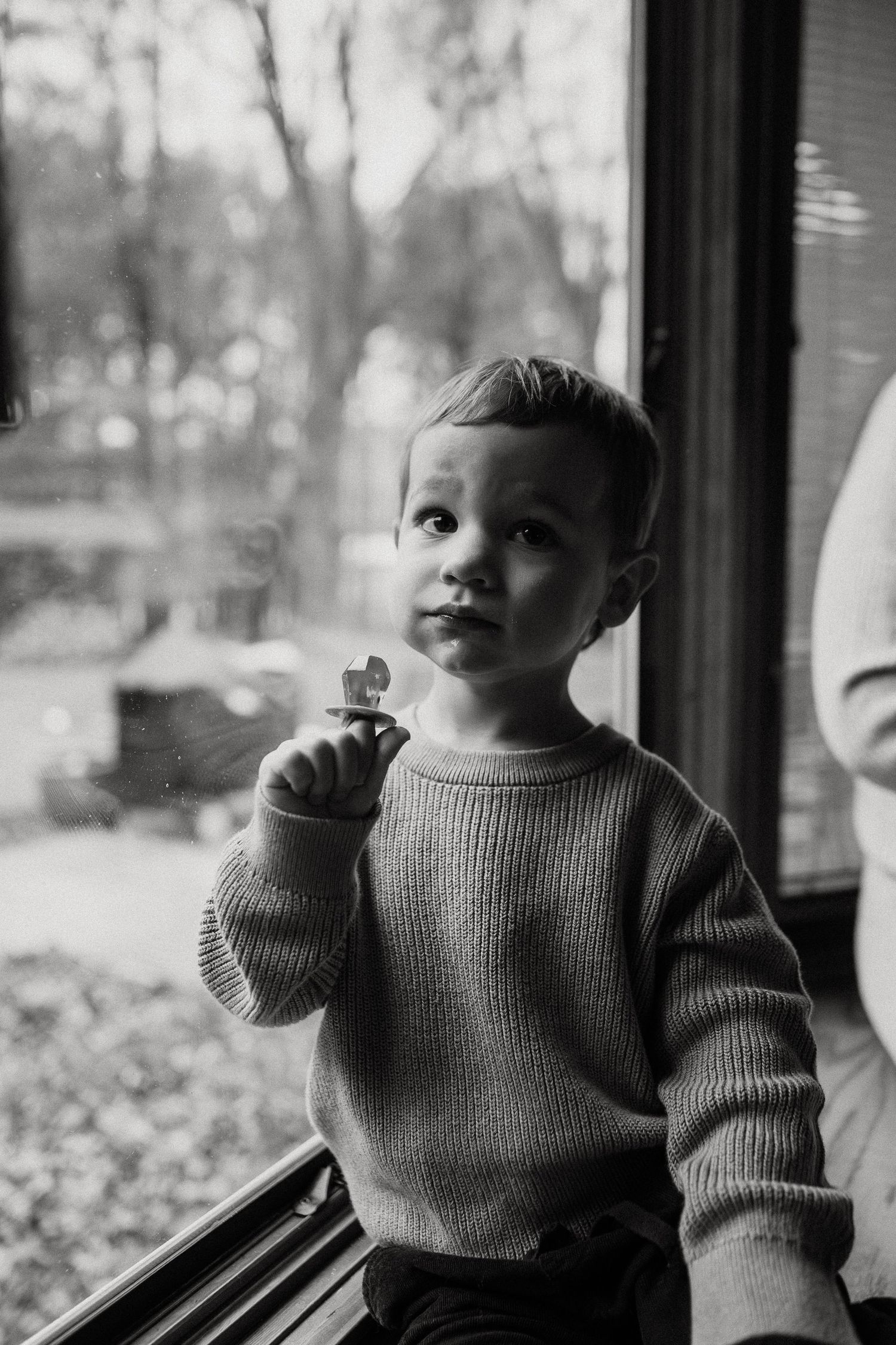 Black and white portrait of baby in knit sweater looking curiously by window.