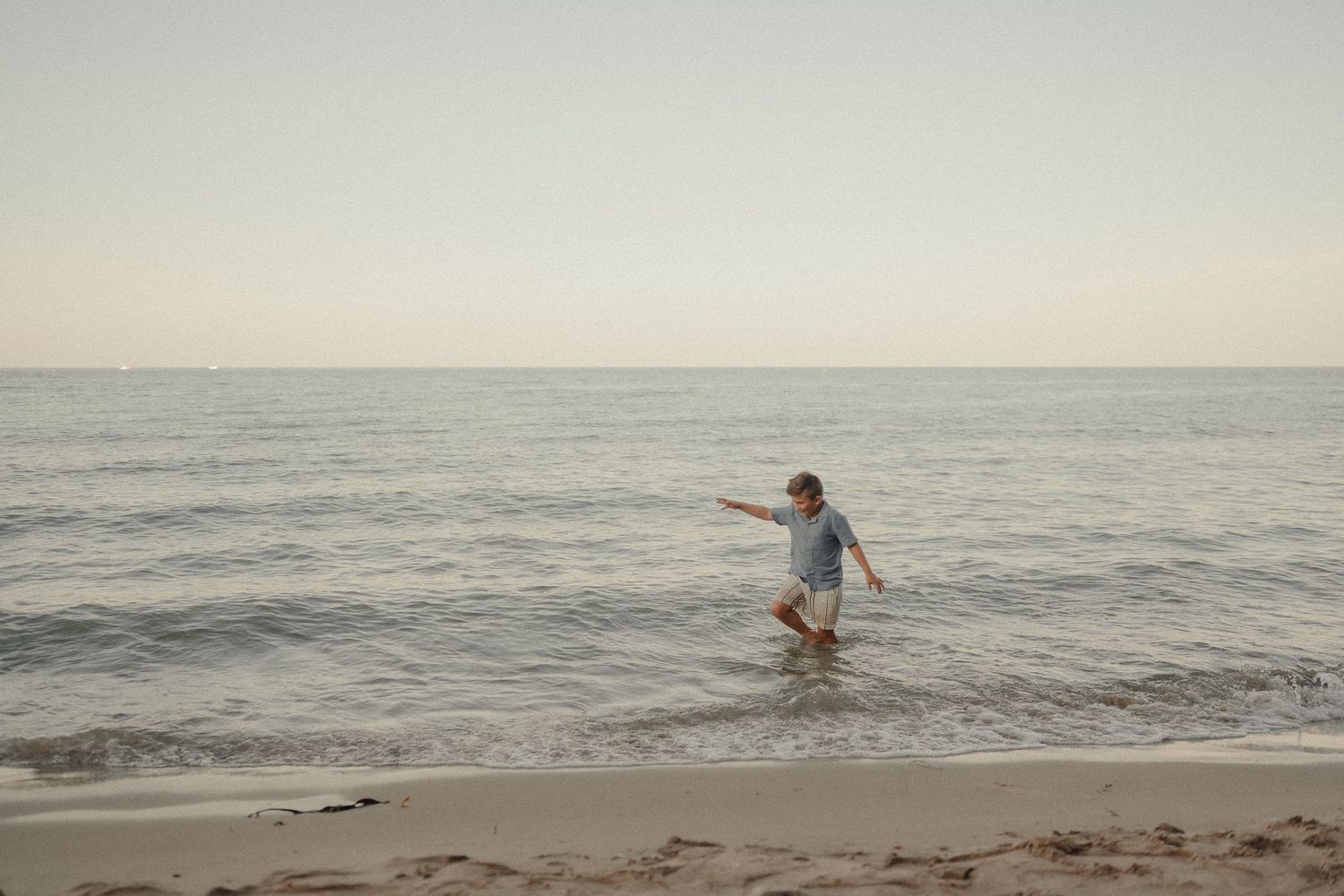 Child wading in ocean waves on a sandy beach at dusk, pointing toward the horizon.