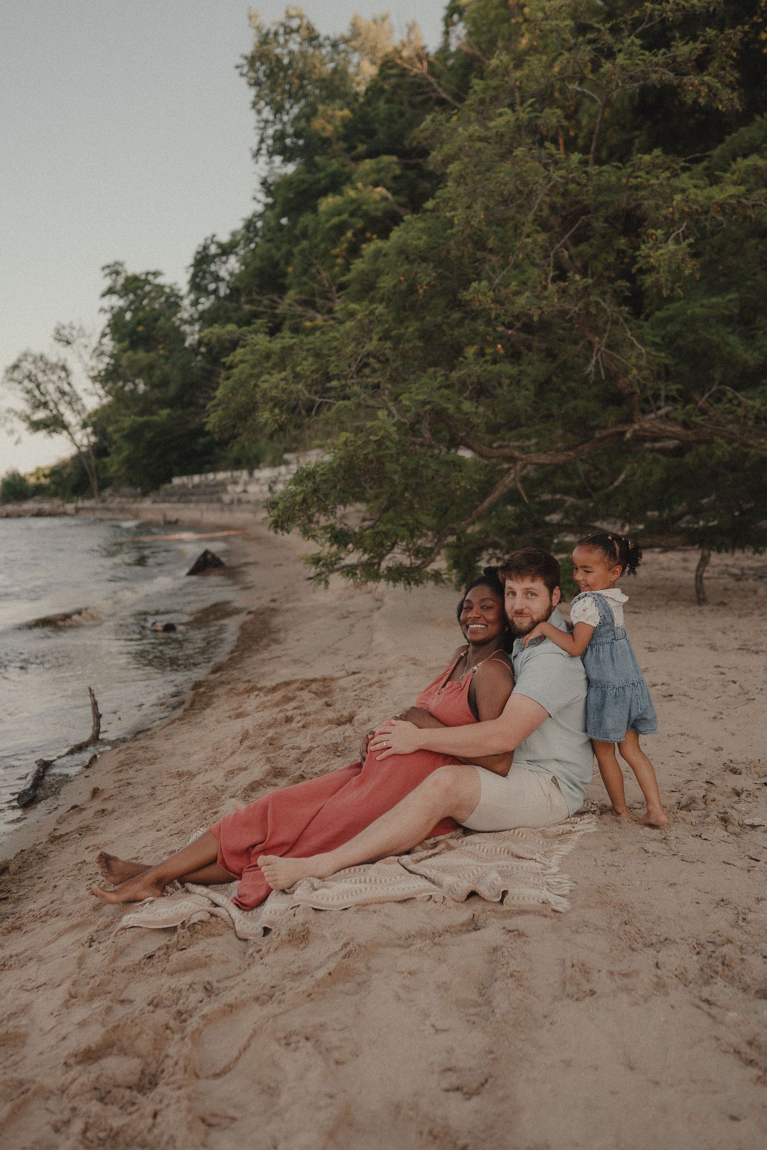A family of three sits on a sandy beach at sunset, with lush green trees in the background.