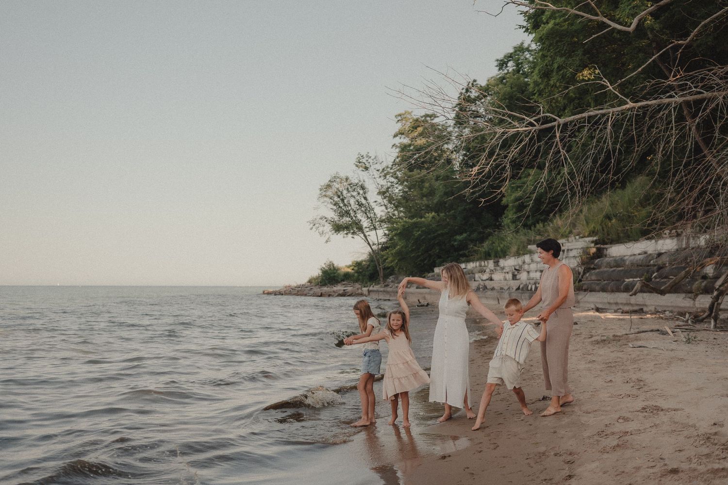 A joyful family dancing and playing together on a sandy beach near calm water with trees in the background.