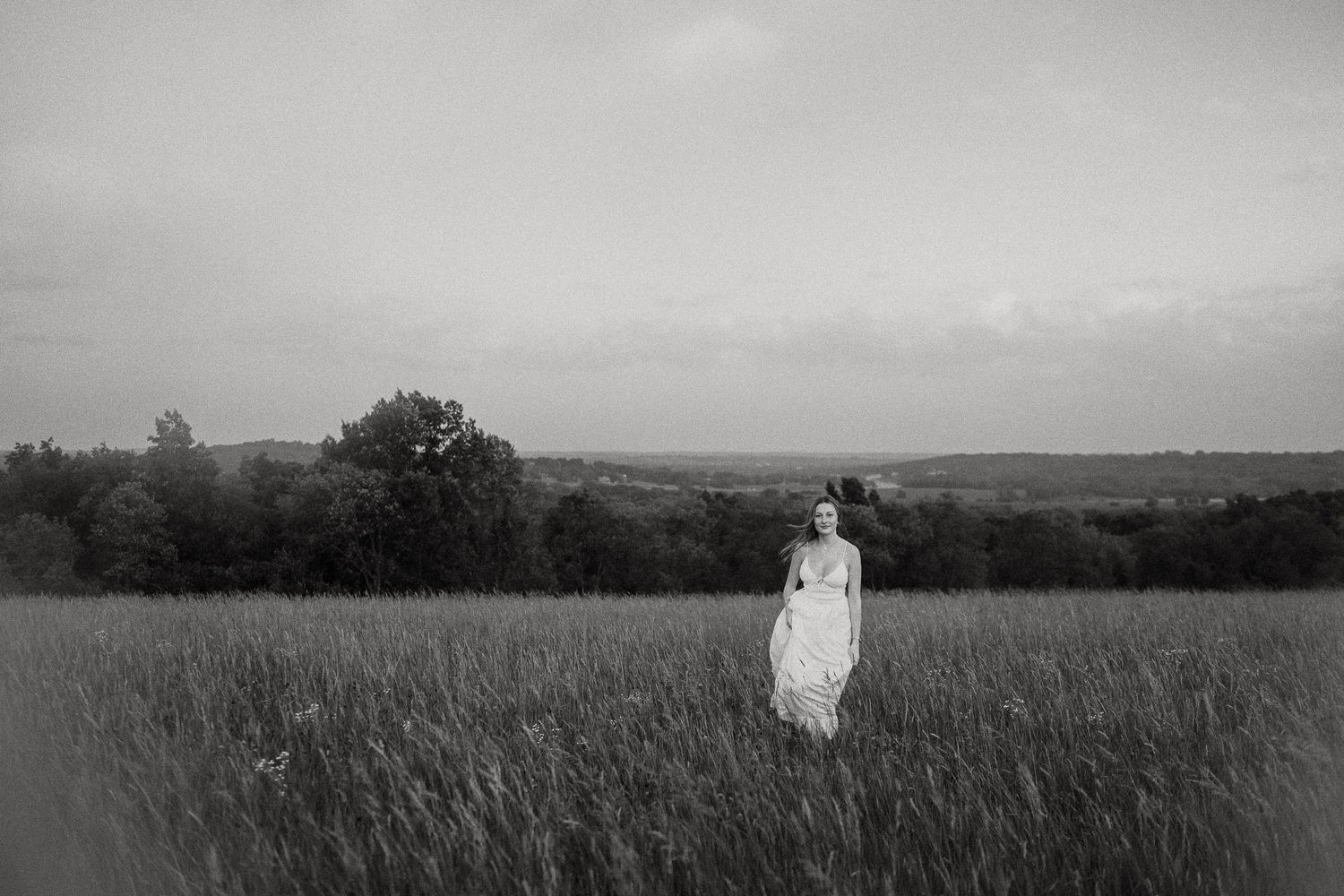 Black and white photo of young woman in white dress standing alone in vast open field.