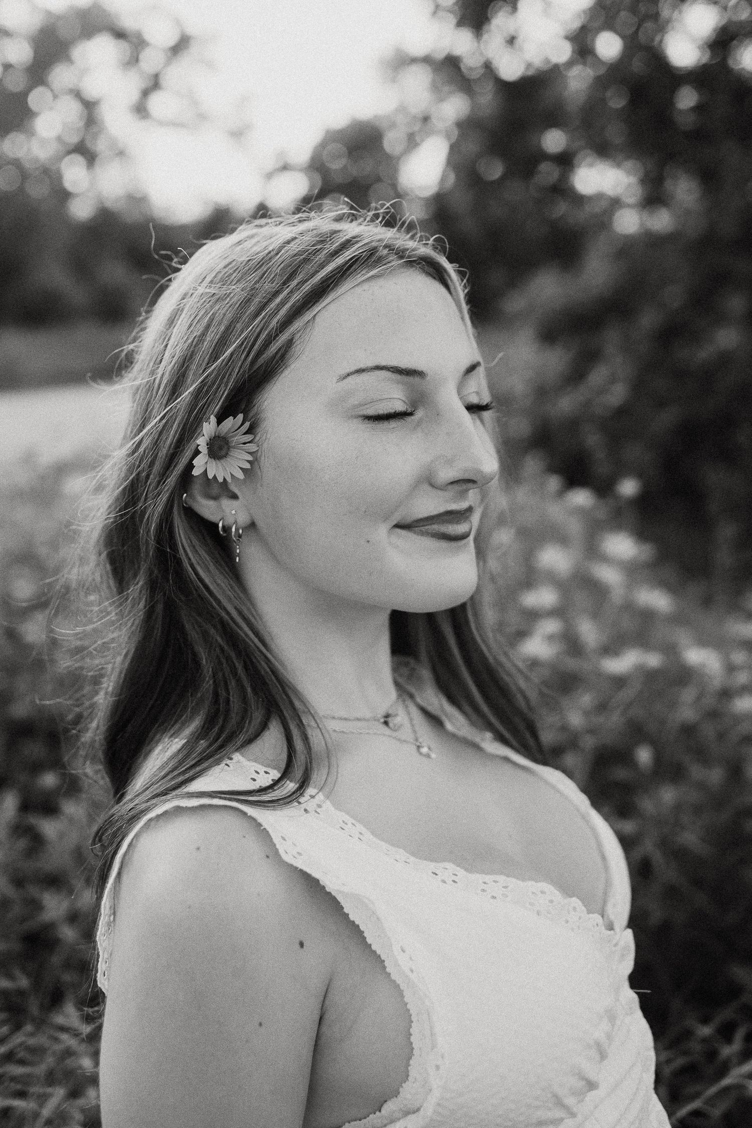 Black and white portrait of a young woman with a daisy in her hair, eyes closed, smiling softly outdoors.