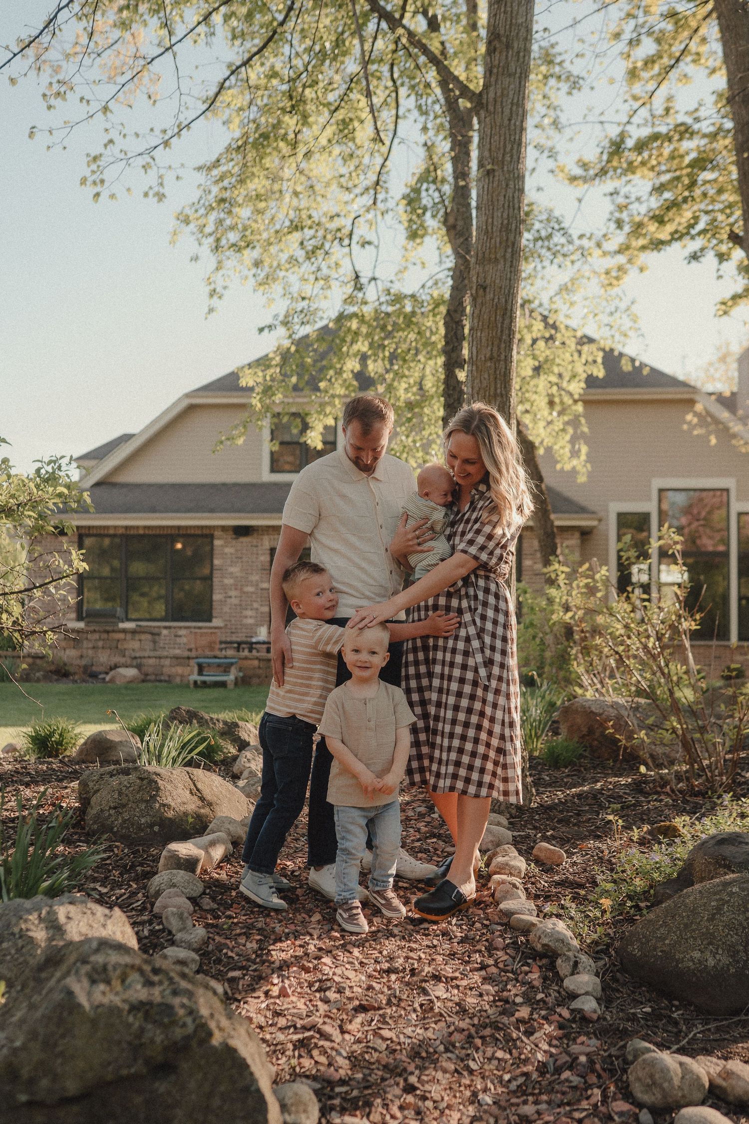 Family of four with newborn posing on a garden path beside large trees in front of a suburban home.