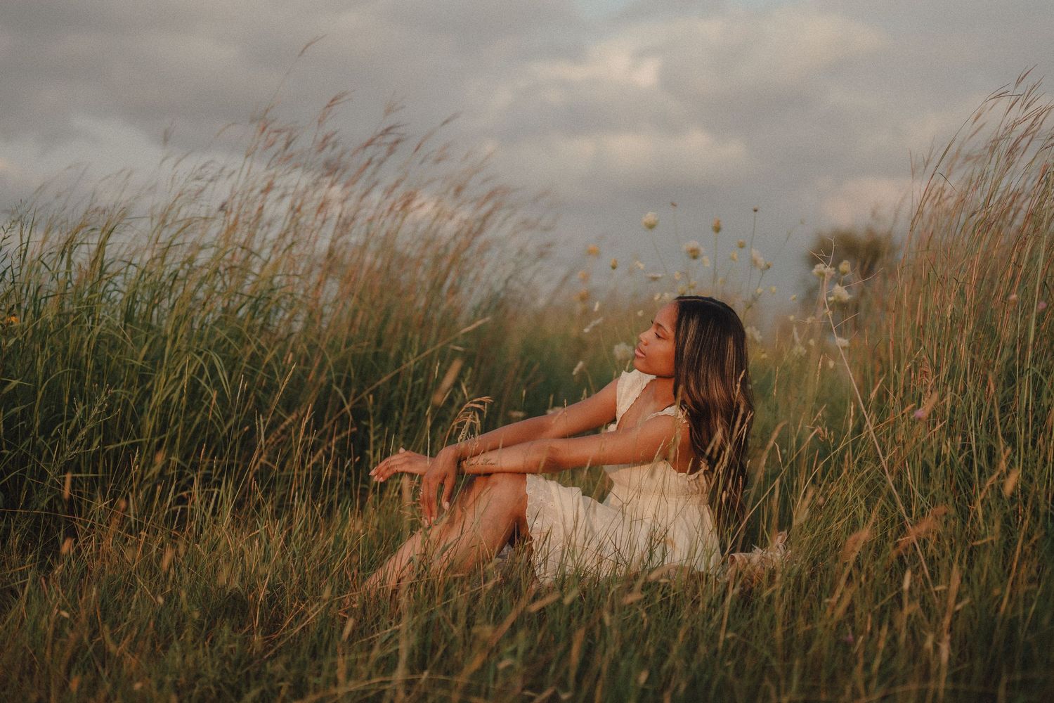 Young woman in a white dress sitting serenely in tall golden grass under a cloudy sky.