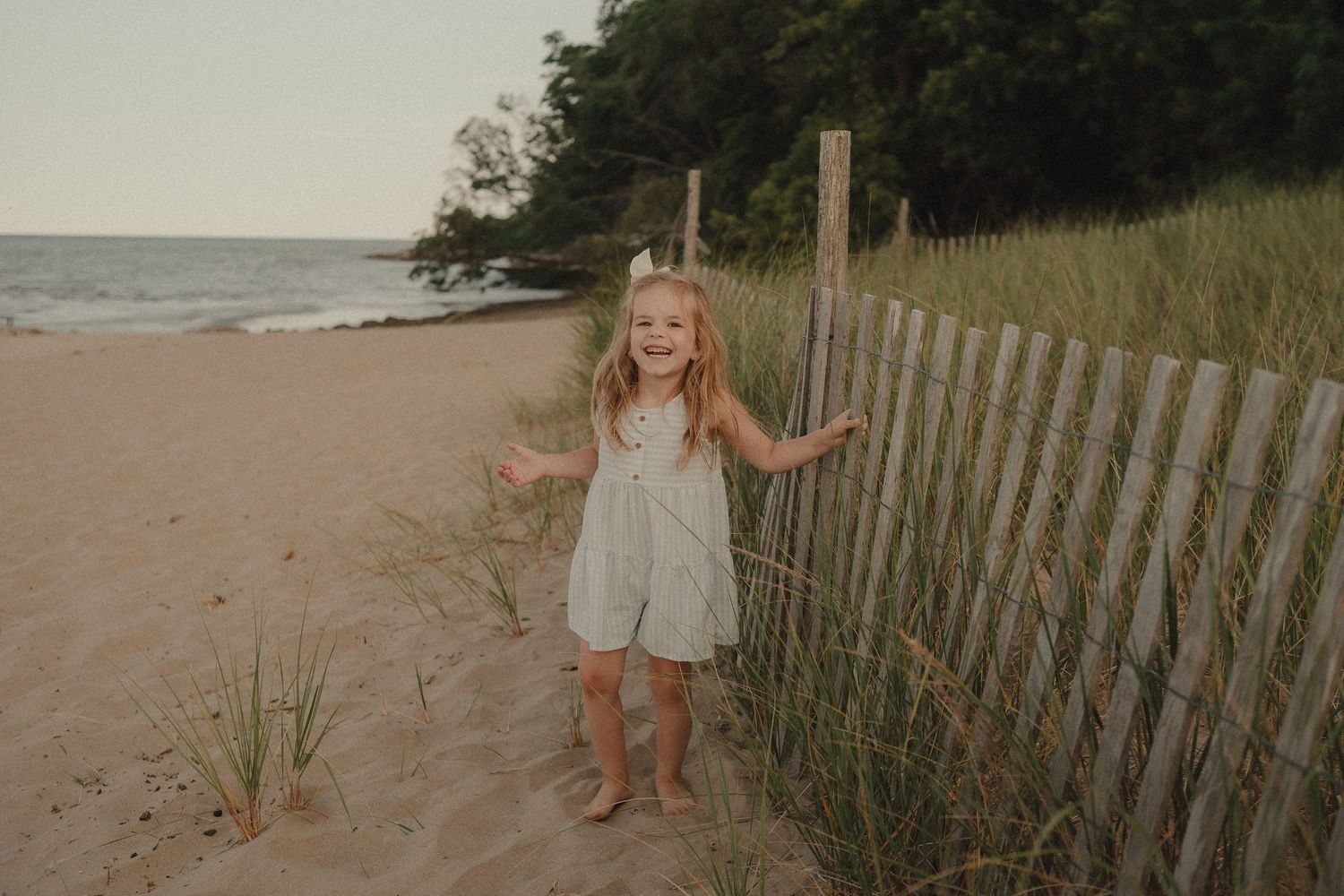 A young girl in a sage romper smiles barefoot on a sandy beach beside a wooden fence at golden hour.