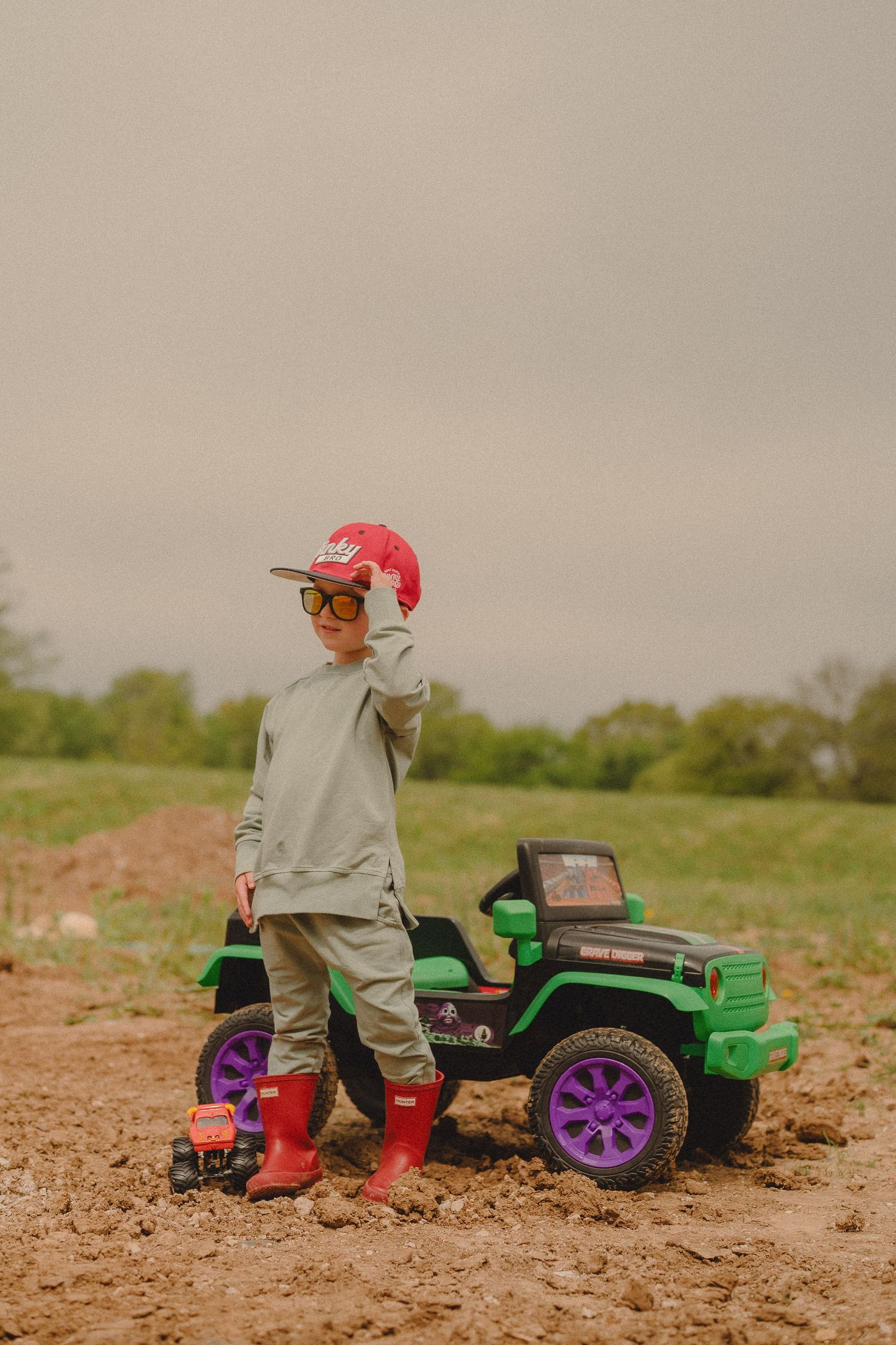 Young boy in red cap and boots posing beside green toy jeep in muddy field.