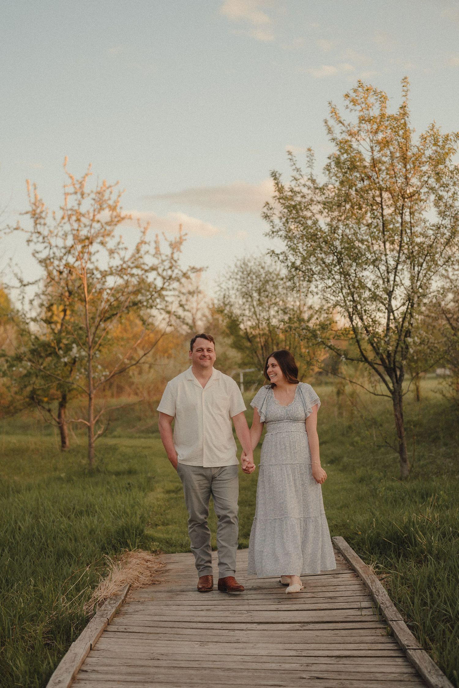 Couple holds hands walking on wooden boardwalk path surrounded by blooming trees at golden hour.