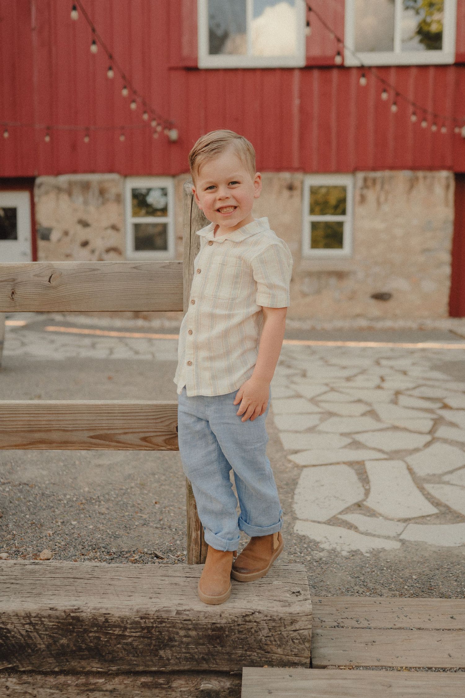Smiling young boy in plaid shirt and jeans posing on wooden steps near a red barn.