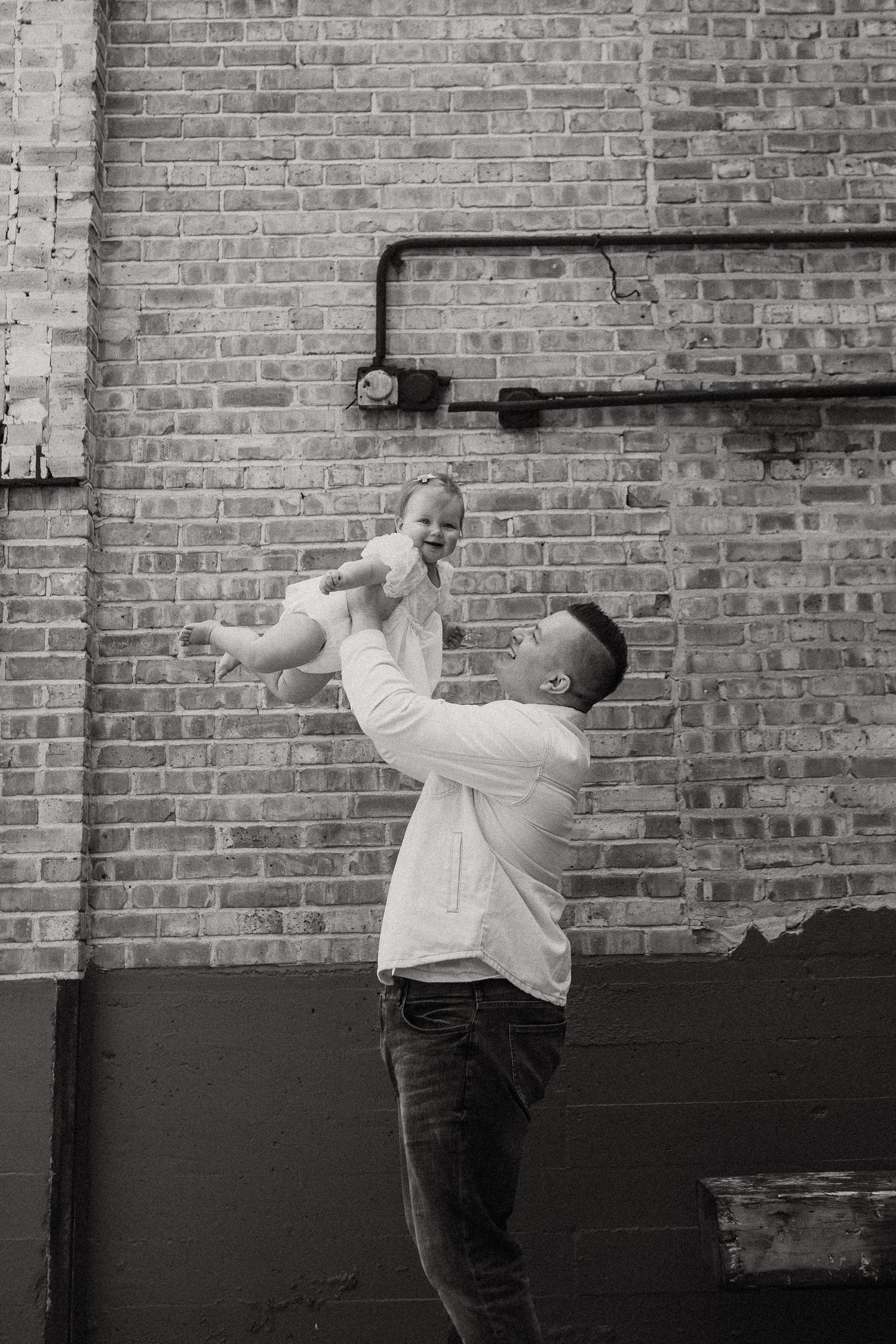 A black and white photo of a father joyfully lifting his laughing baby in the air outdoors.