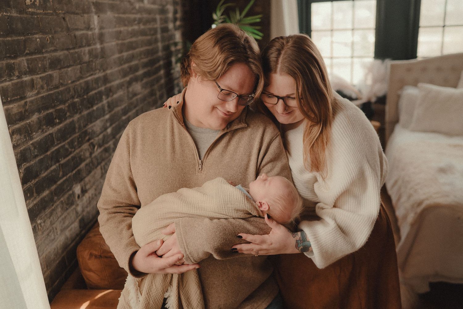 Two new parents in glasses tenderly hold their swaddled newborn baby in a cozy brick-walled bedroom.