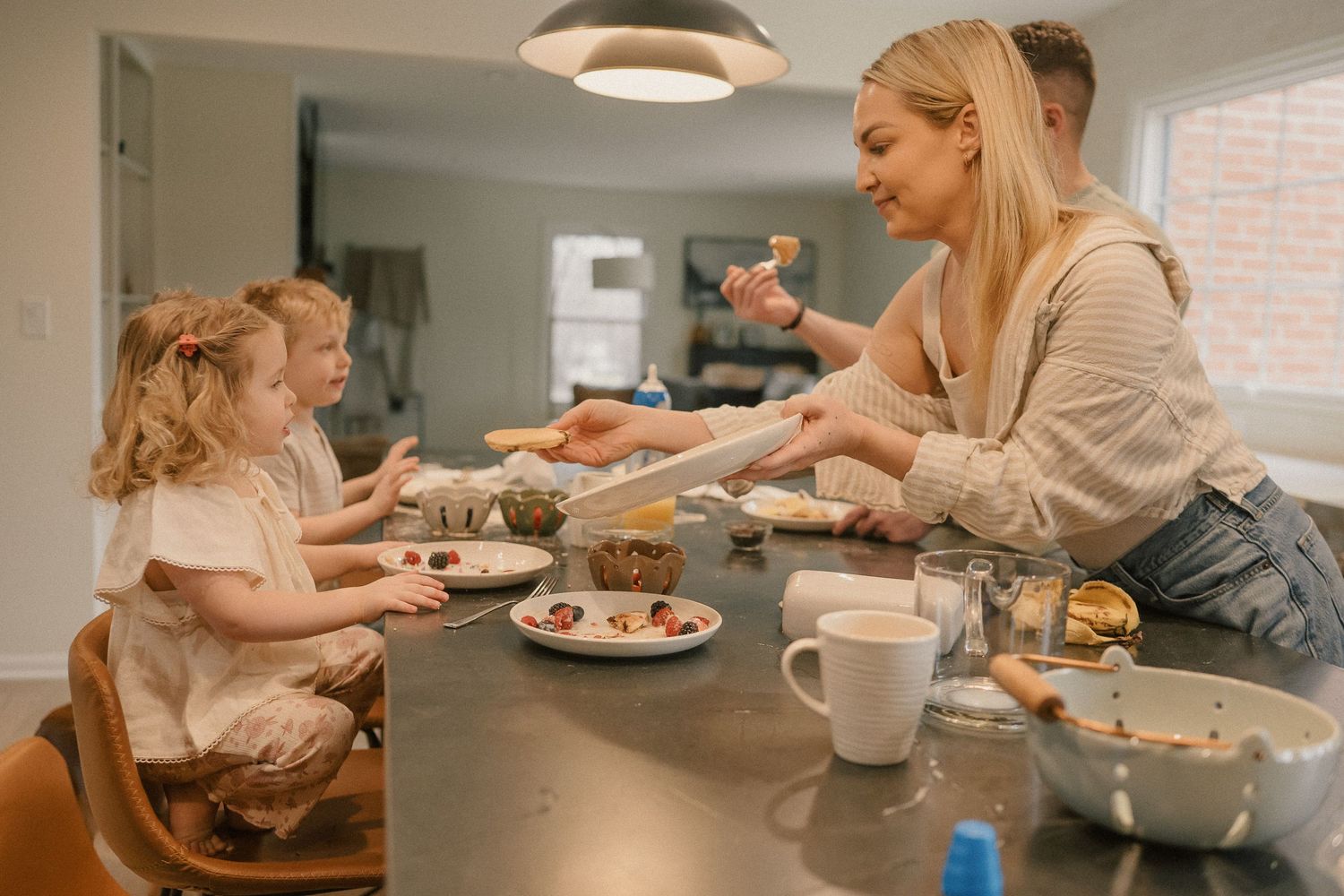 A mother serves food to her two young children at a dark kitchen counter with a mug nearby.