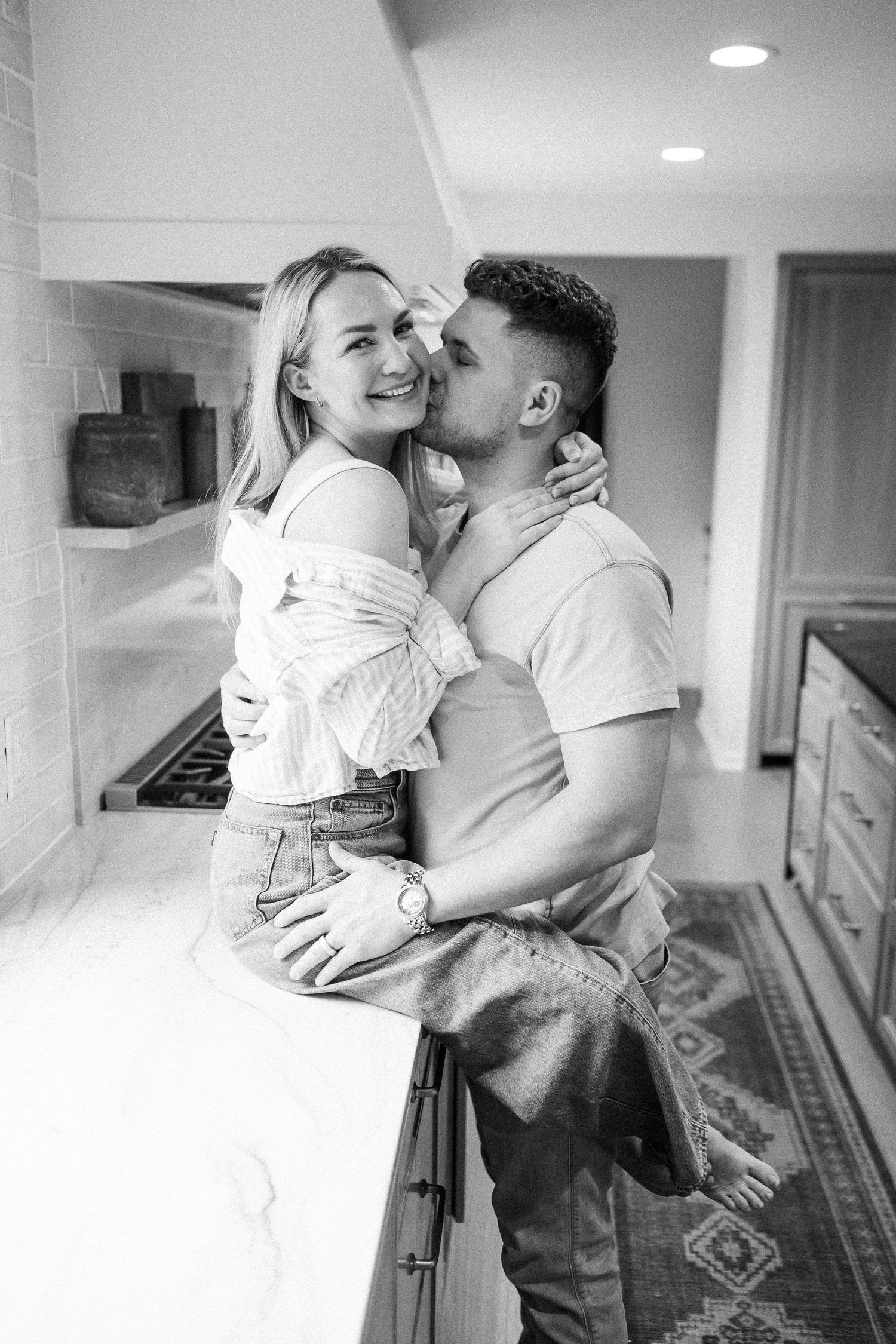 Black and white photo of a couple embracing in a modern kitchen, man kissing womans cheek.