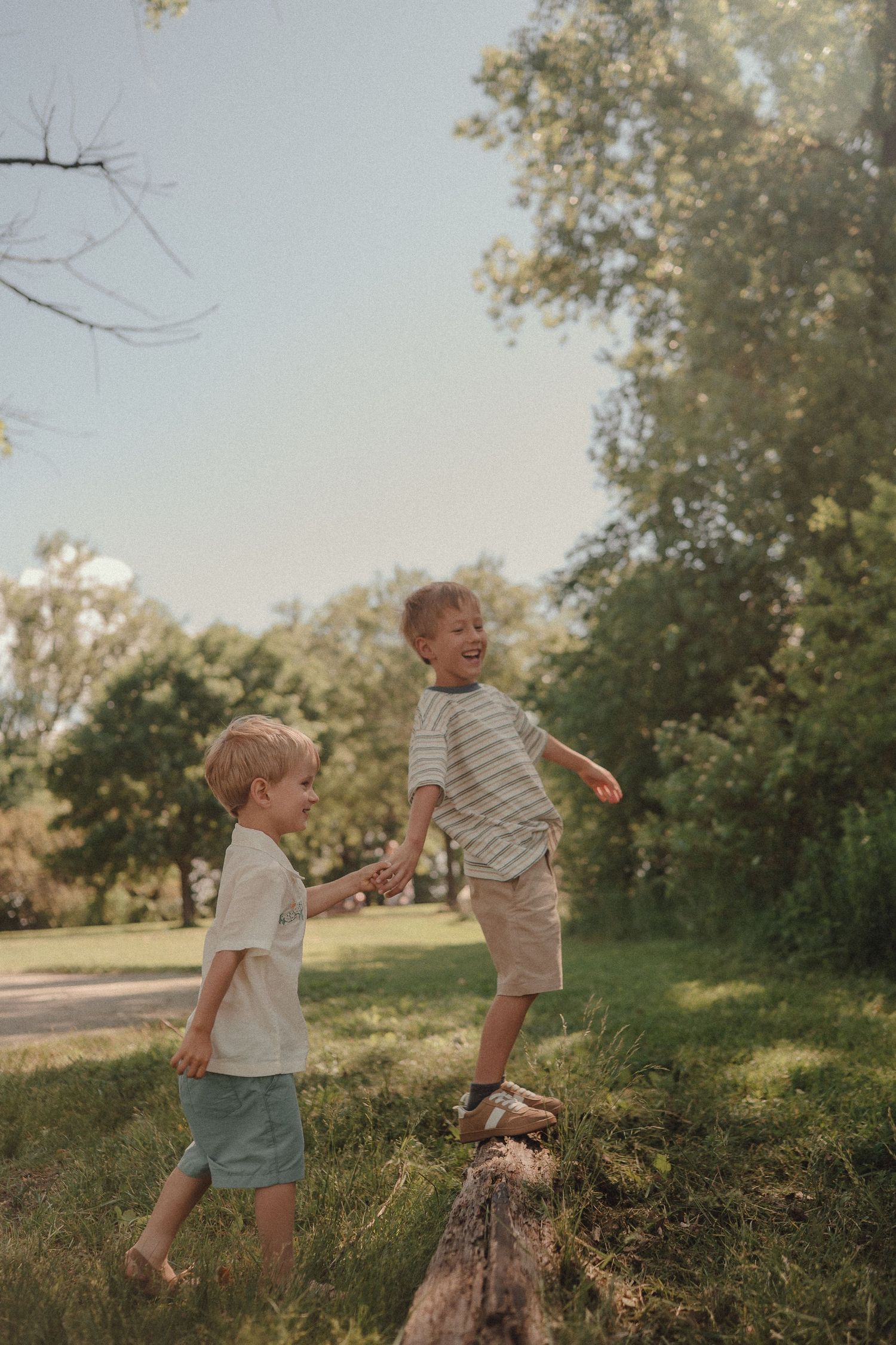 Two young brothers laugh and play on a fallen log in a sunny green park.