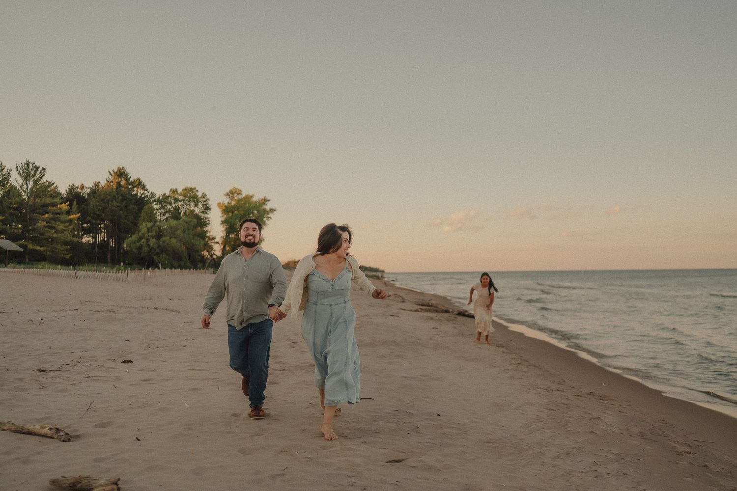 Couple walking hand-in-hand on sandy beach at dusk with child playing near ocean waves.