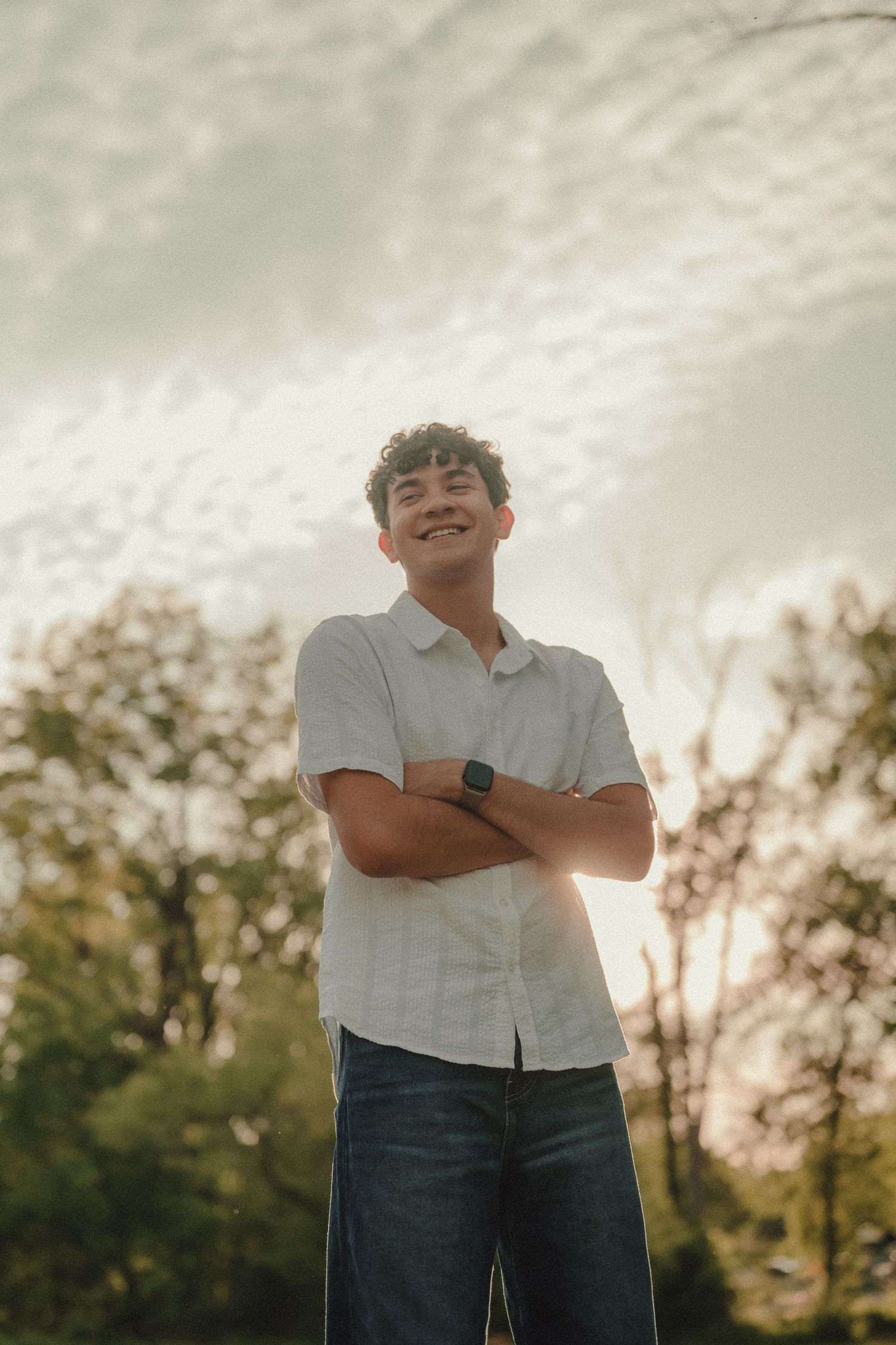 Smiling young man with curly hair, arms crossed, wearing a white shirt and smartwatch, standing outdoors near trees.