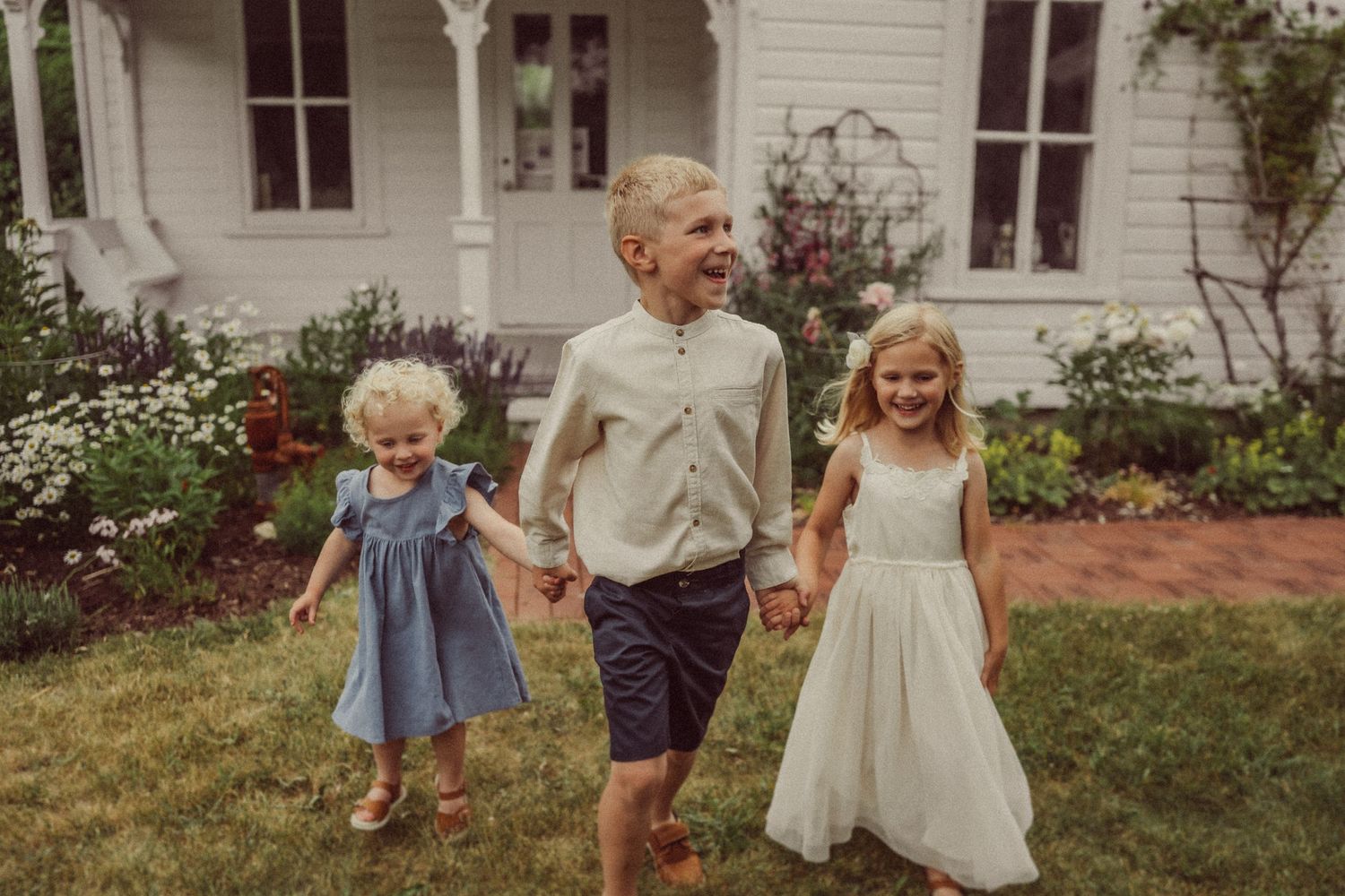 Children holding hands while walking through garden by white farmhouse.