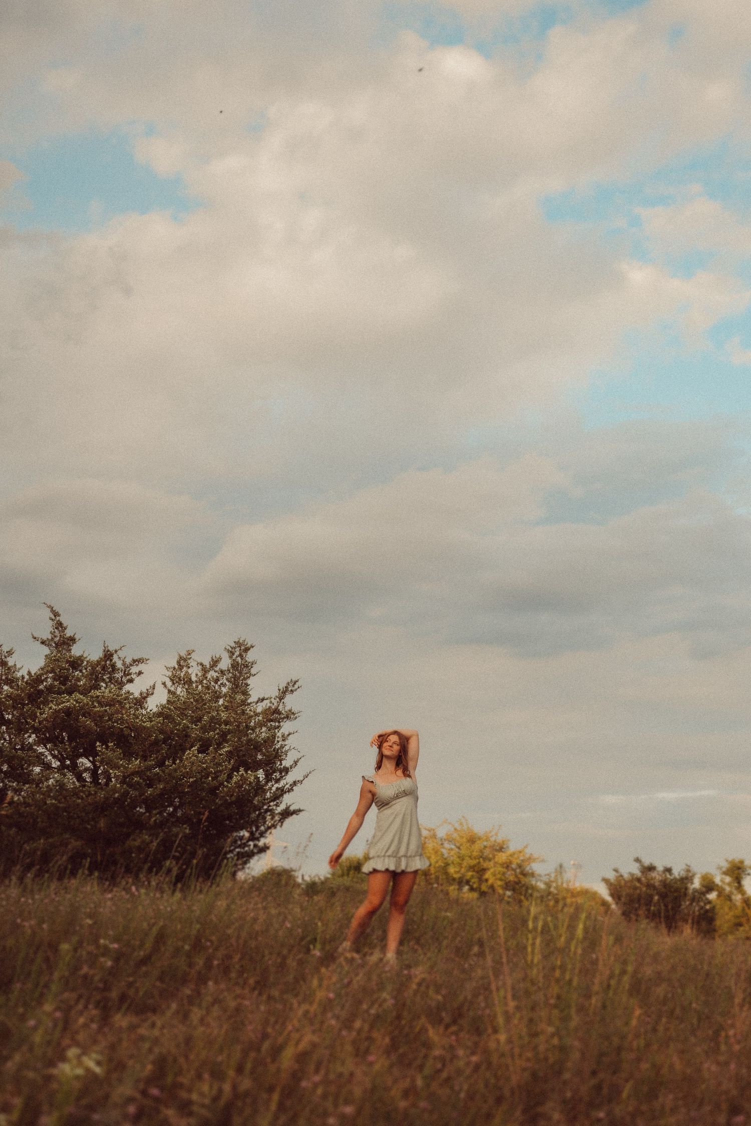 Figure in flowing dress stands on grassy hilltop against dramatic cloudy sky.