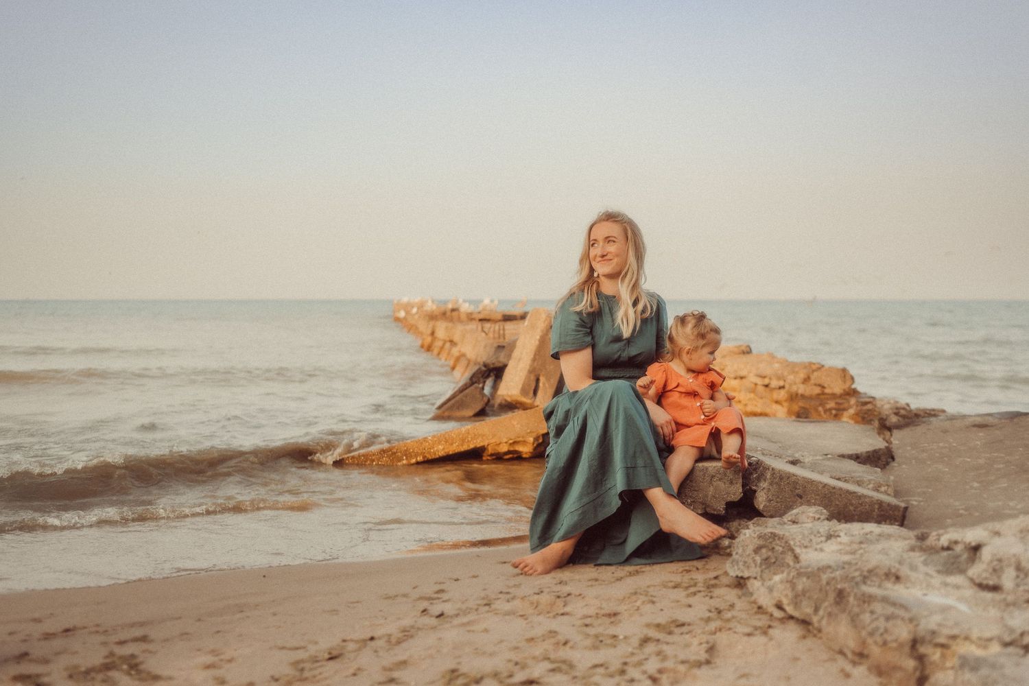 Two people in casual attire sit together on coastal rocks at sunset by the ocean.
