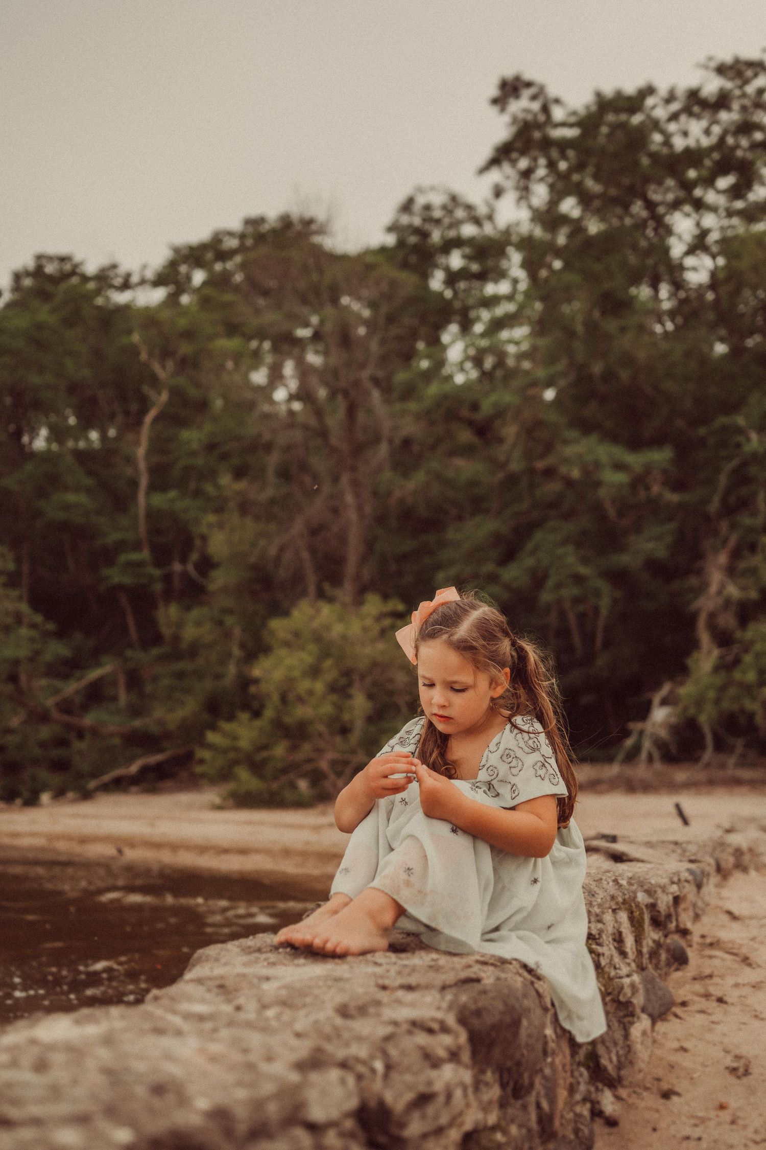 Child in flowing dress sits thoughtfully on coastal rocks near wooded shoreline.