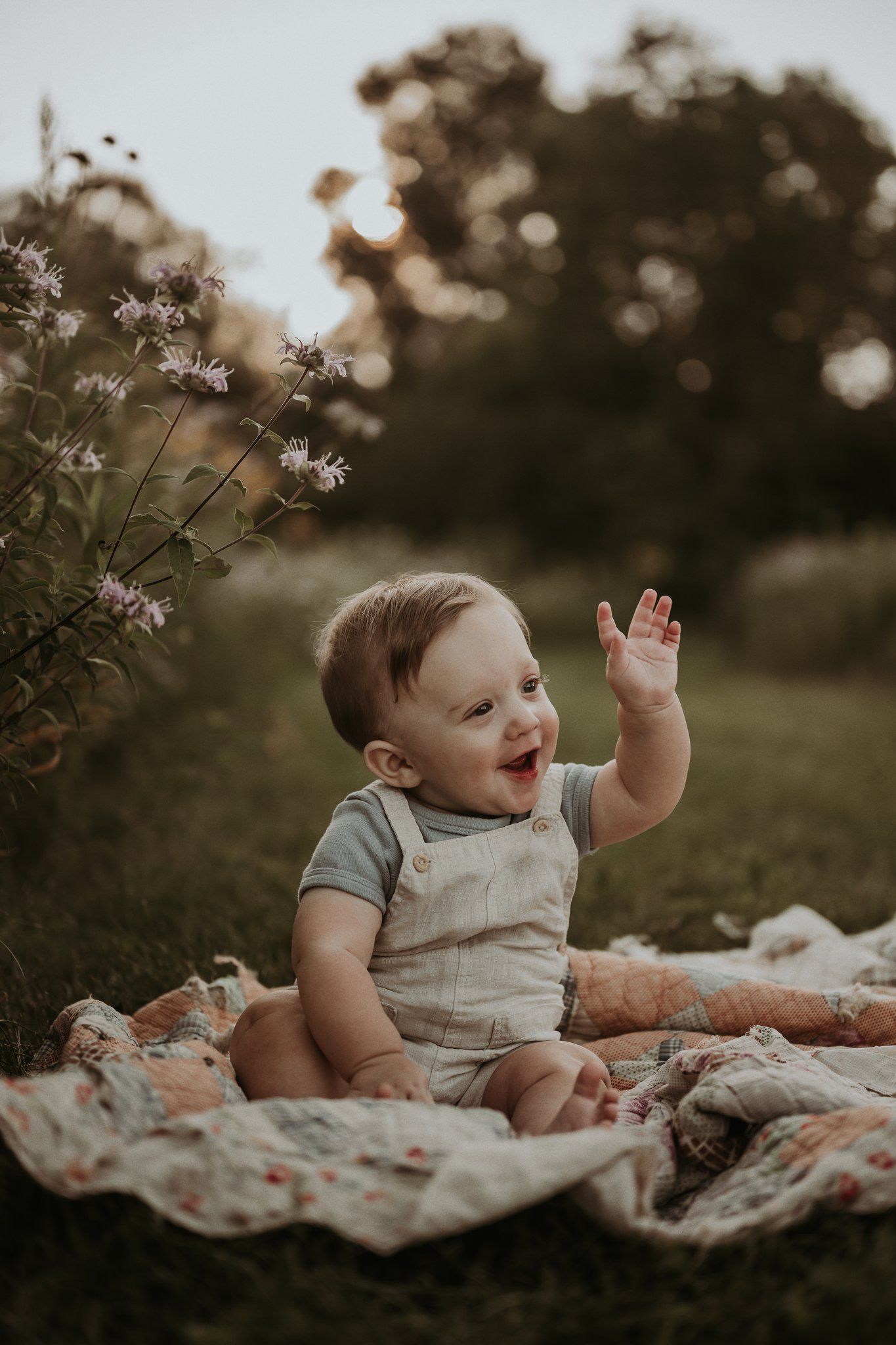 Happy baby in overalls sits on blanket outdoors reaching toward wildflowers.