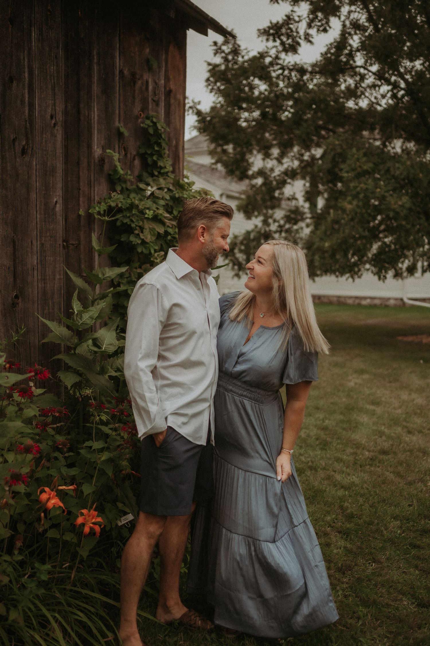 Romantic couple in coordinated outfits standing by a rustic barn with flowers blooming nearby.