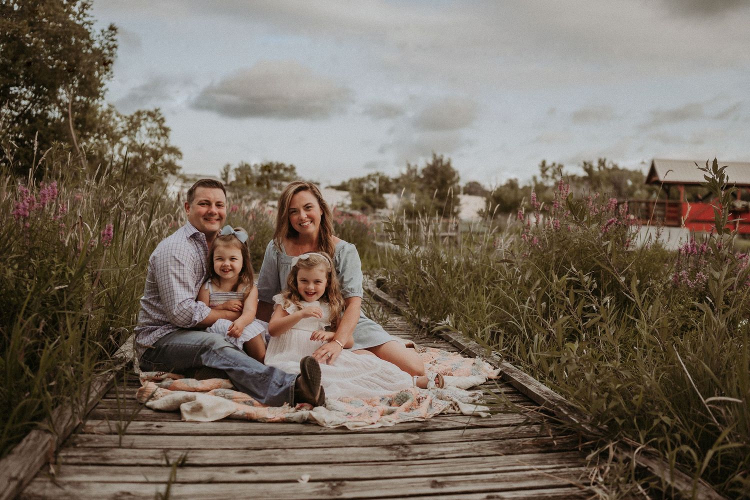 Relaxed family portrait on wooden boardwalk surrounded by wild grasses at sunset.
